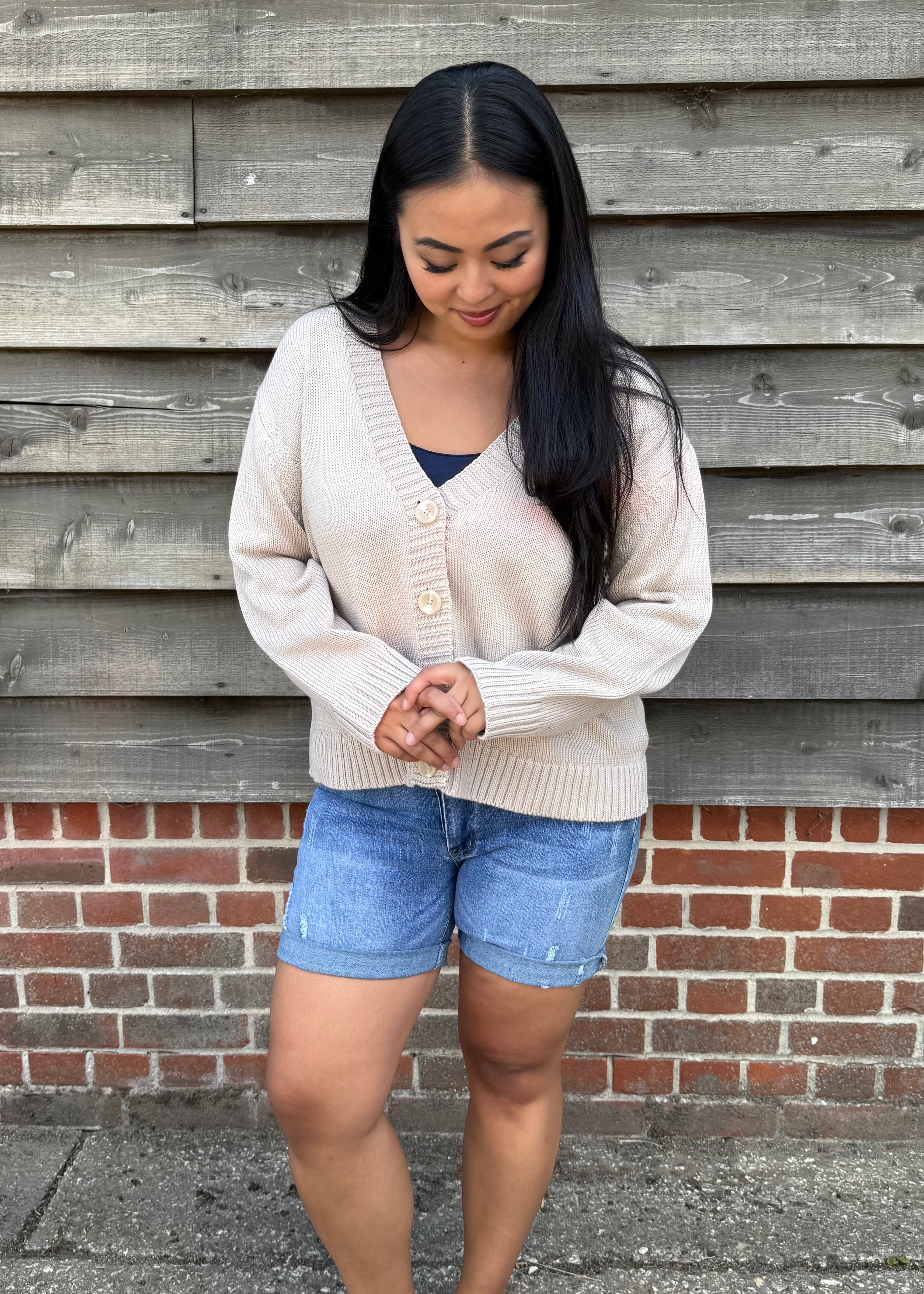 Woman wearing a beige cardigan and denim shorts standing against a wooden wall.