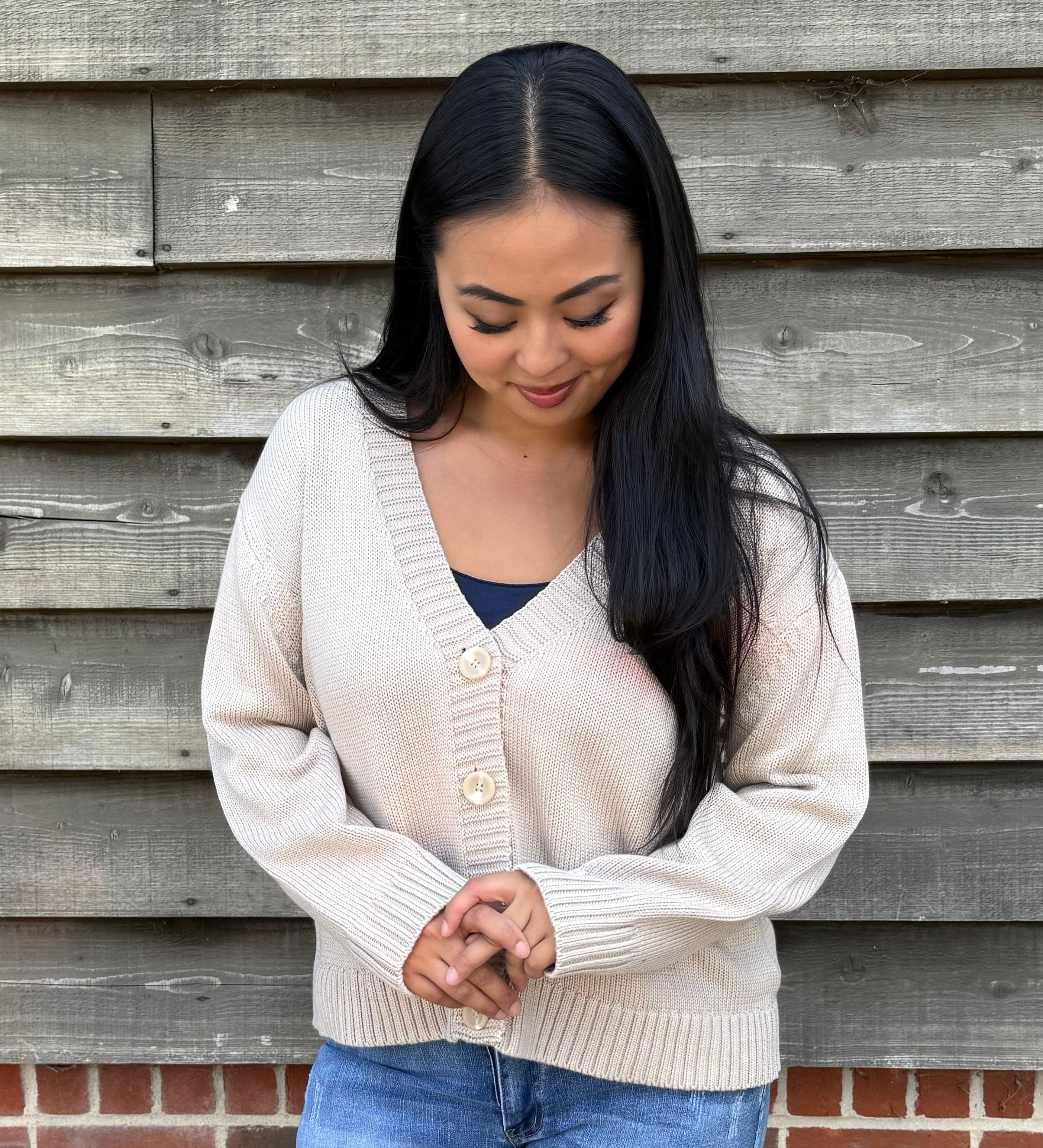 Woman wearing a beige cardigan and denim shorts standing against a wooden wall.