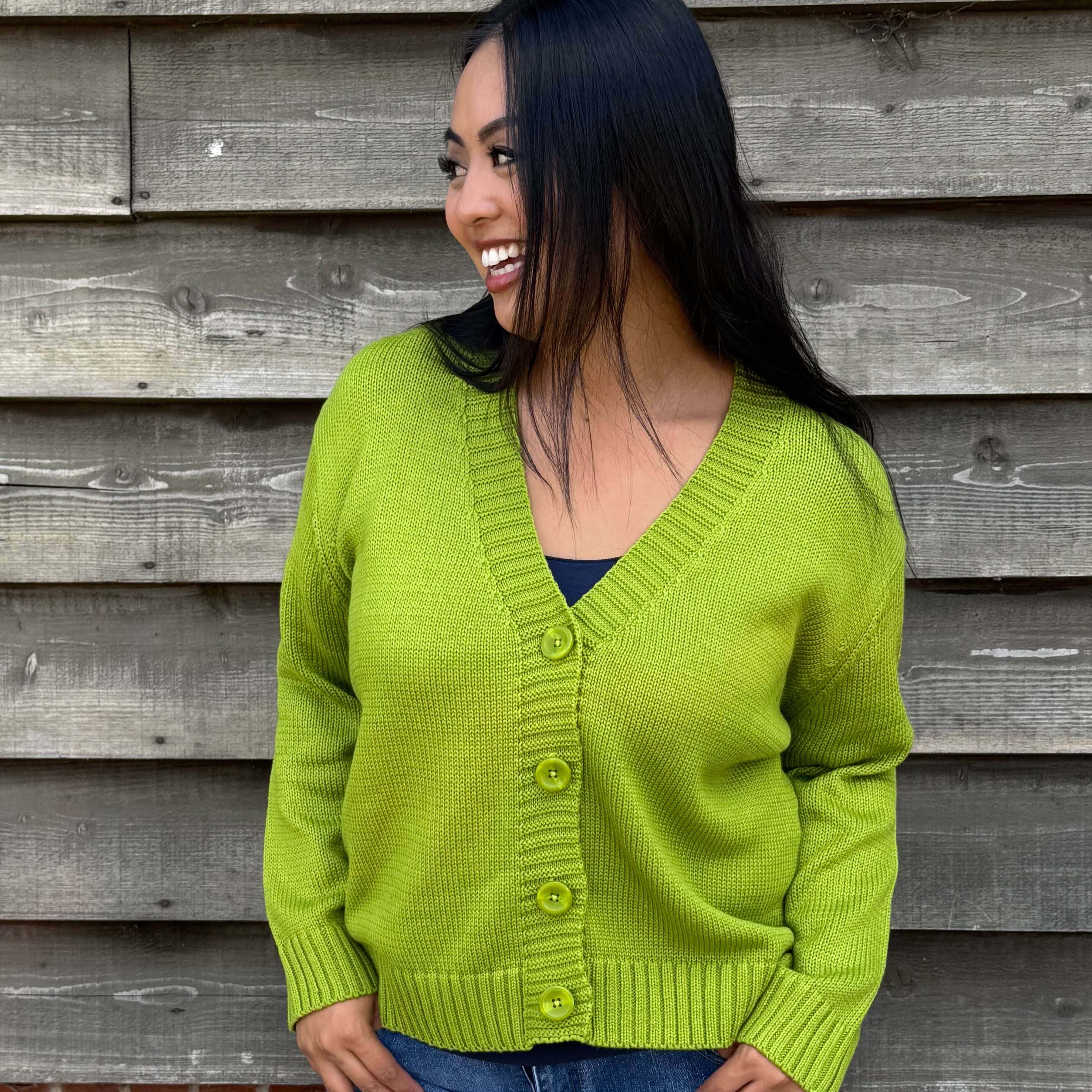Woman wearing a green cardigan and denim shorts standing against a wooden wall.