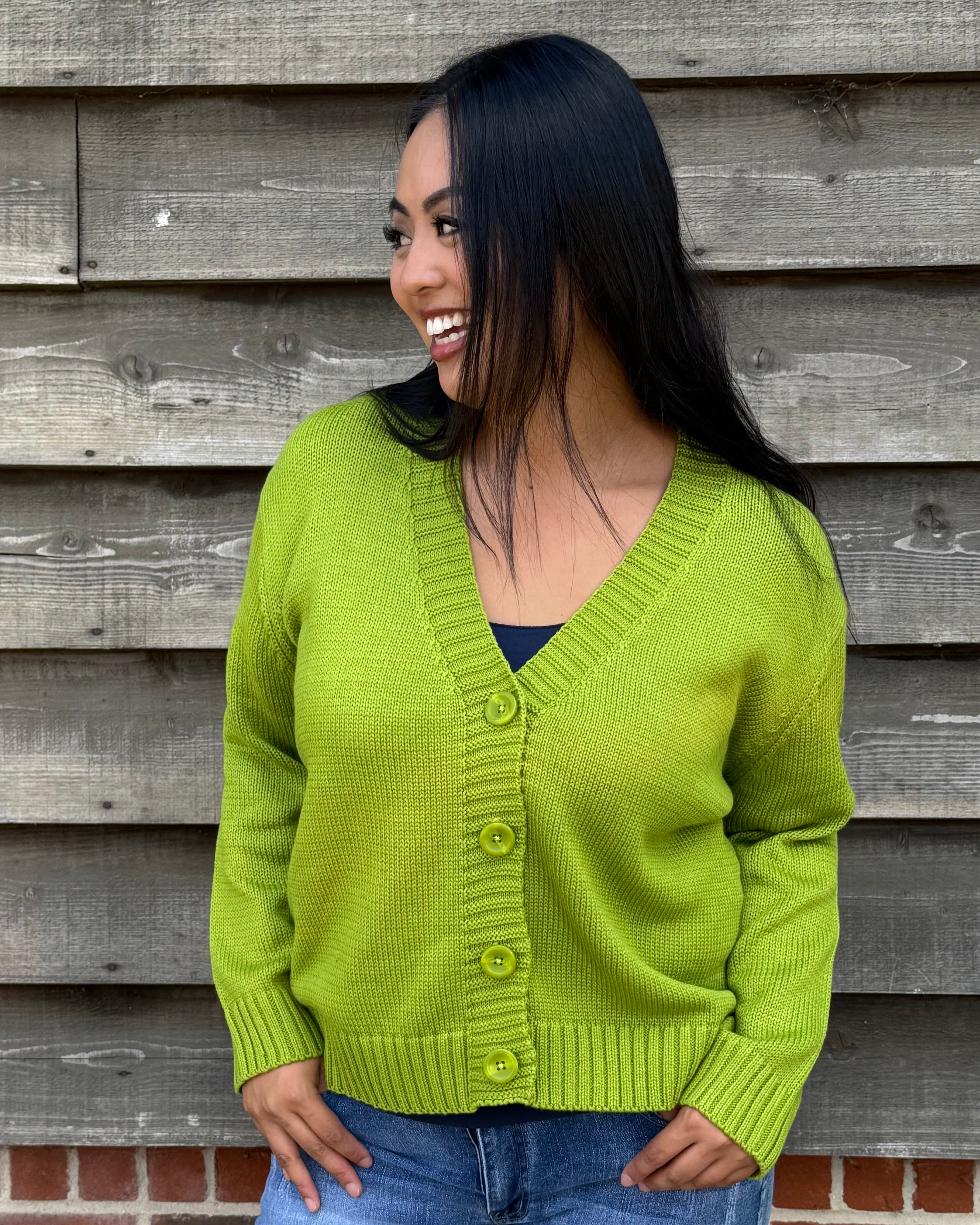 Woman wearing a green cardigan and denim shorts standing against a wooden wall.