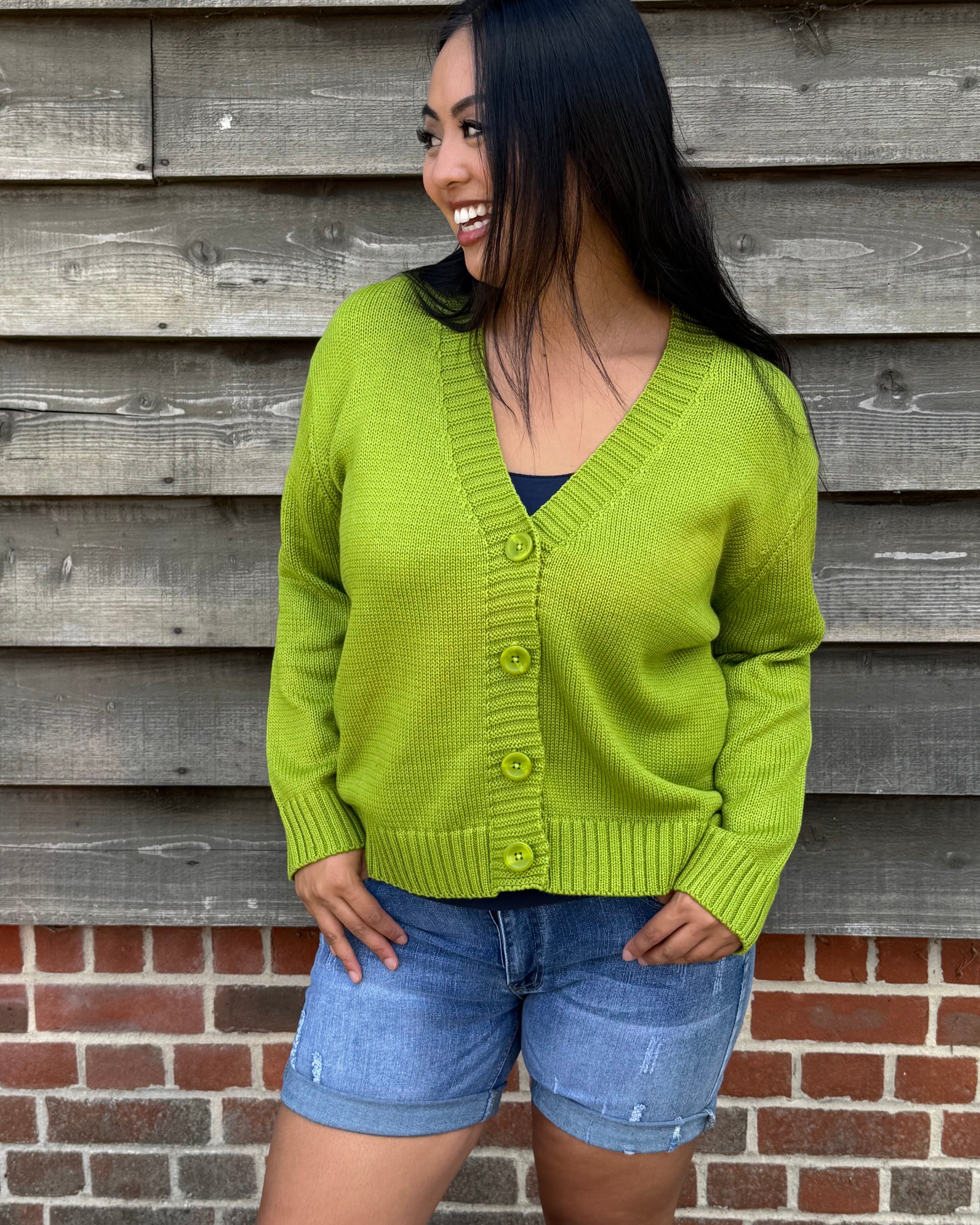 Woman wearing a green cardigan and denim shorts standing against a wooden wall.