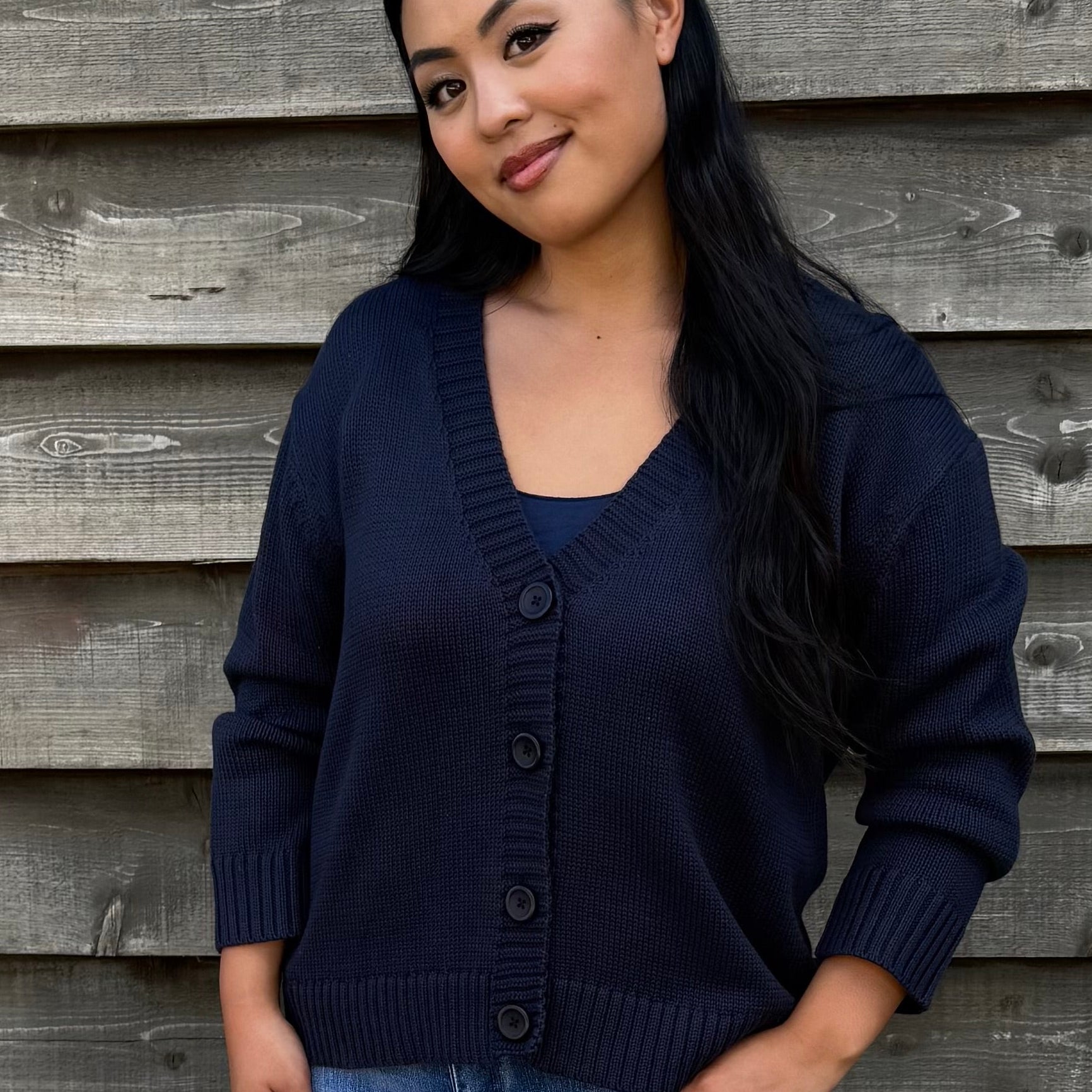 Woman wearing a navy cardigan, denim shorts, and white sandals standing against a wooden wall.