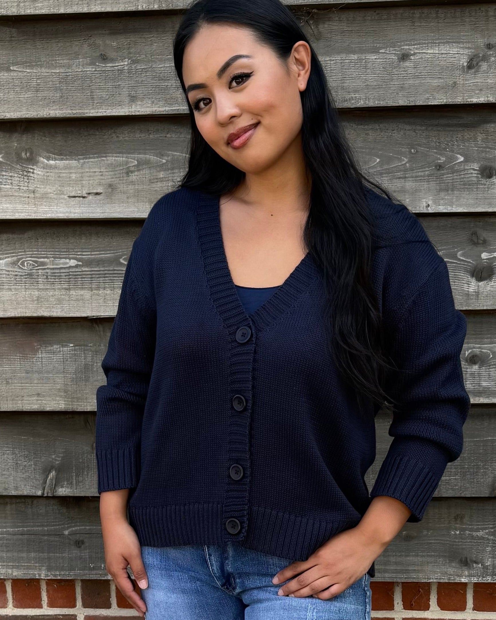 Woman wearing a navy cardigan, denim shorts, and white sandals standing against a wooden wall.