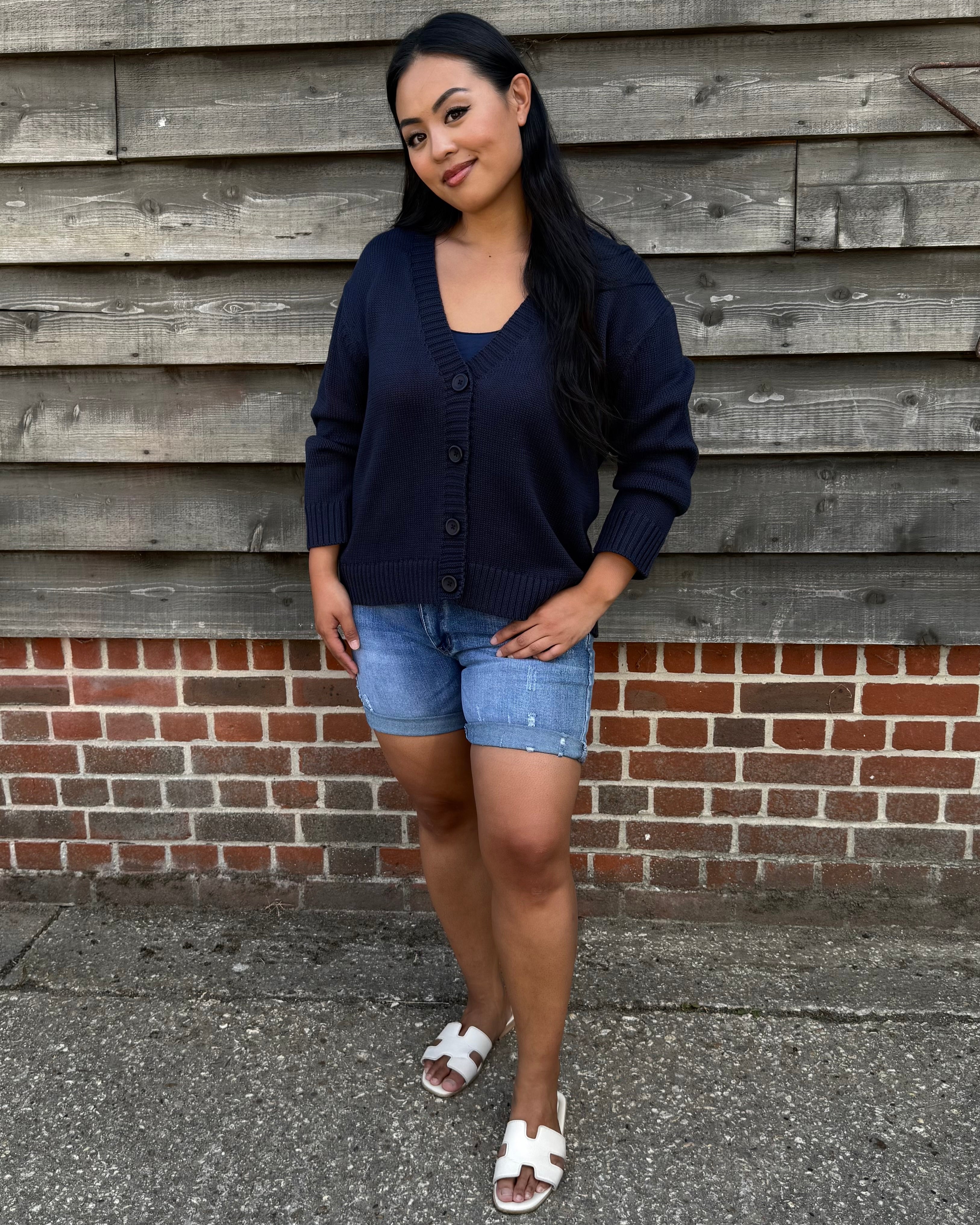 Woman wearing a navy cardigan, denim shorts, and white sandals standing against a wooden wall.