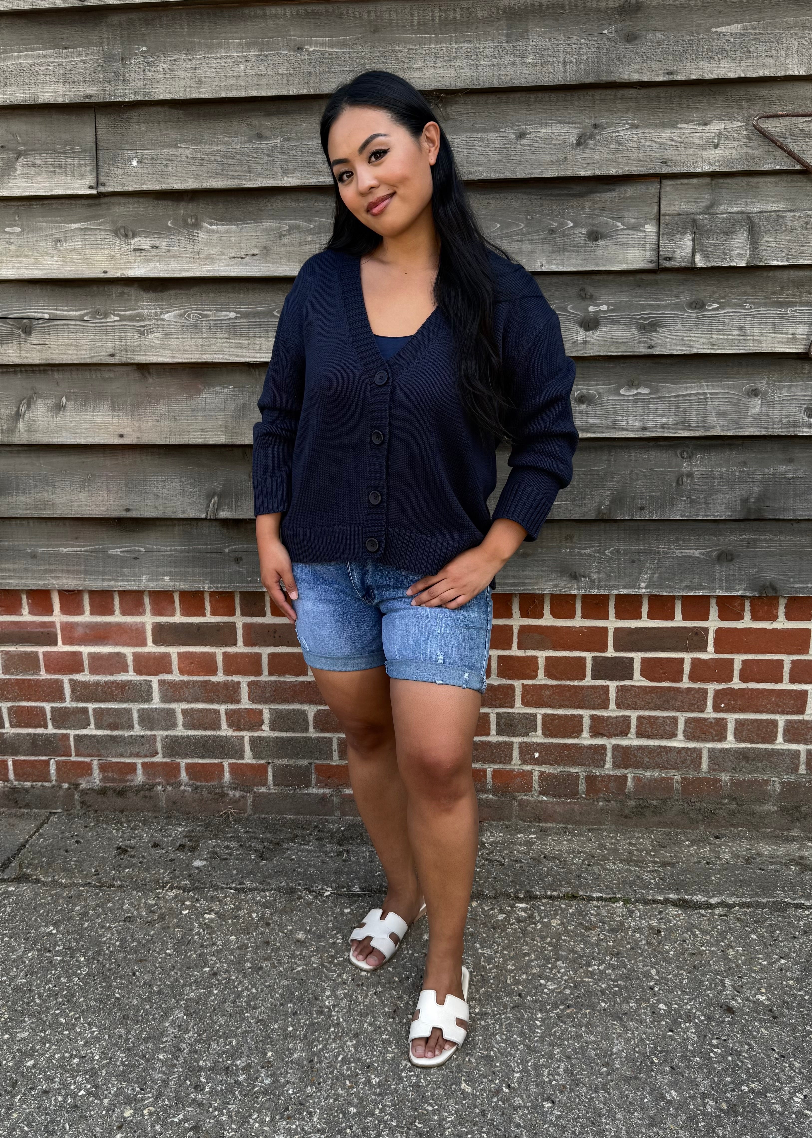 Woman wearing a navy cardigan, denim shorts, and white sandals standing against a wooden wall.