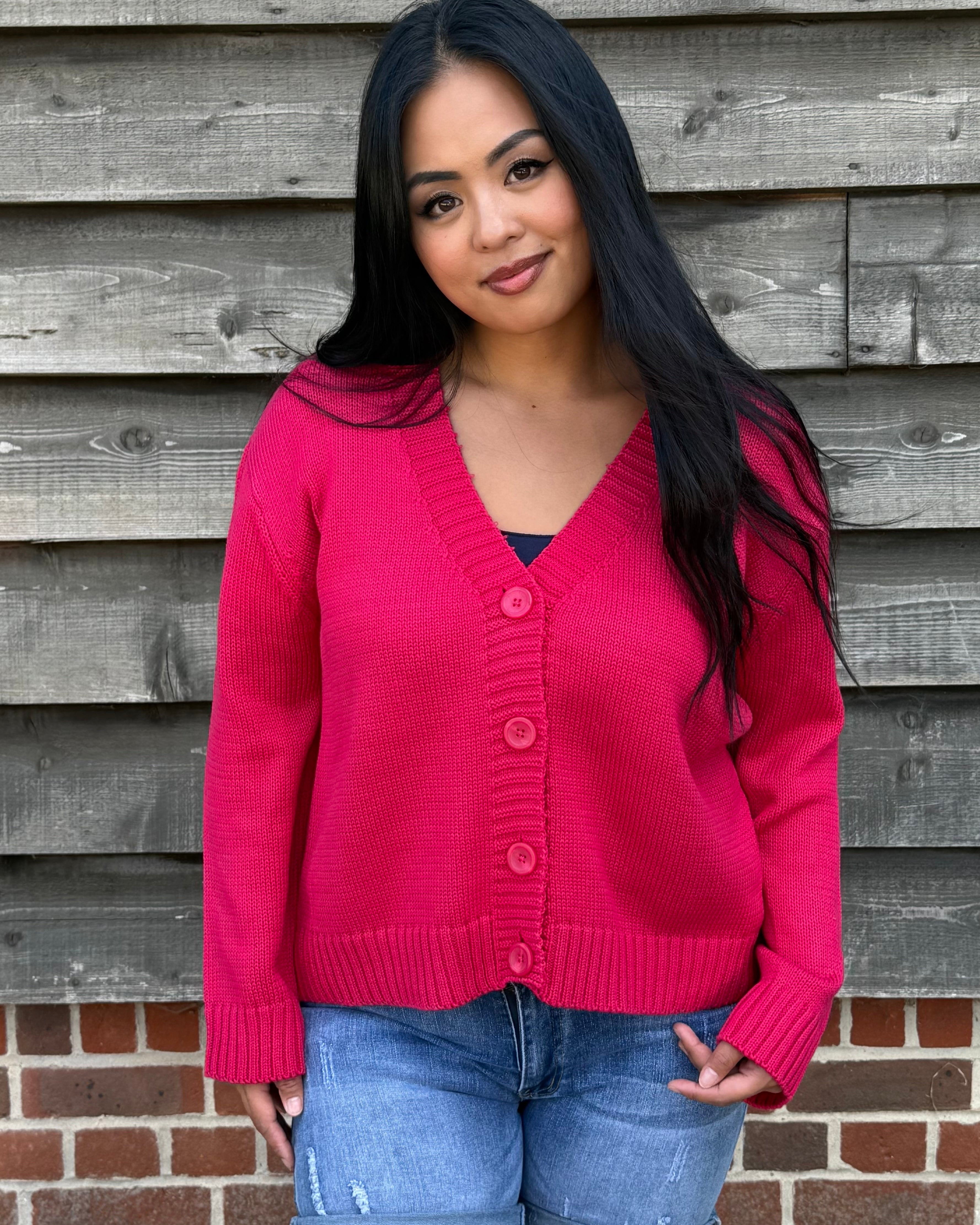 Woman wearing a pink cardigan and denim shorts standing against a wooden wall.