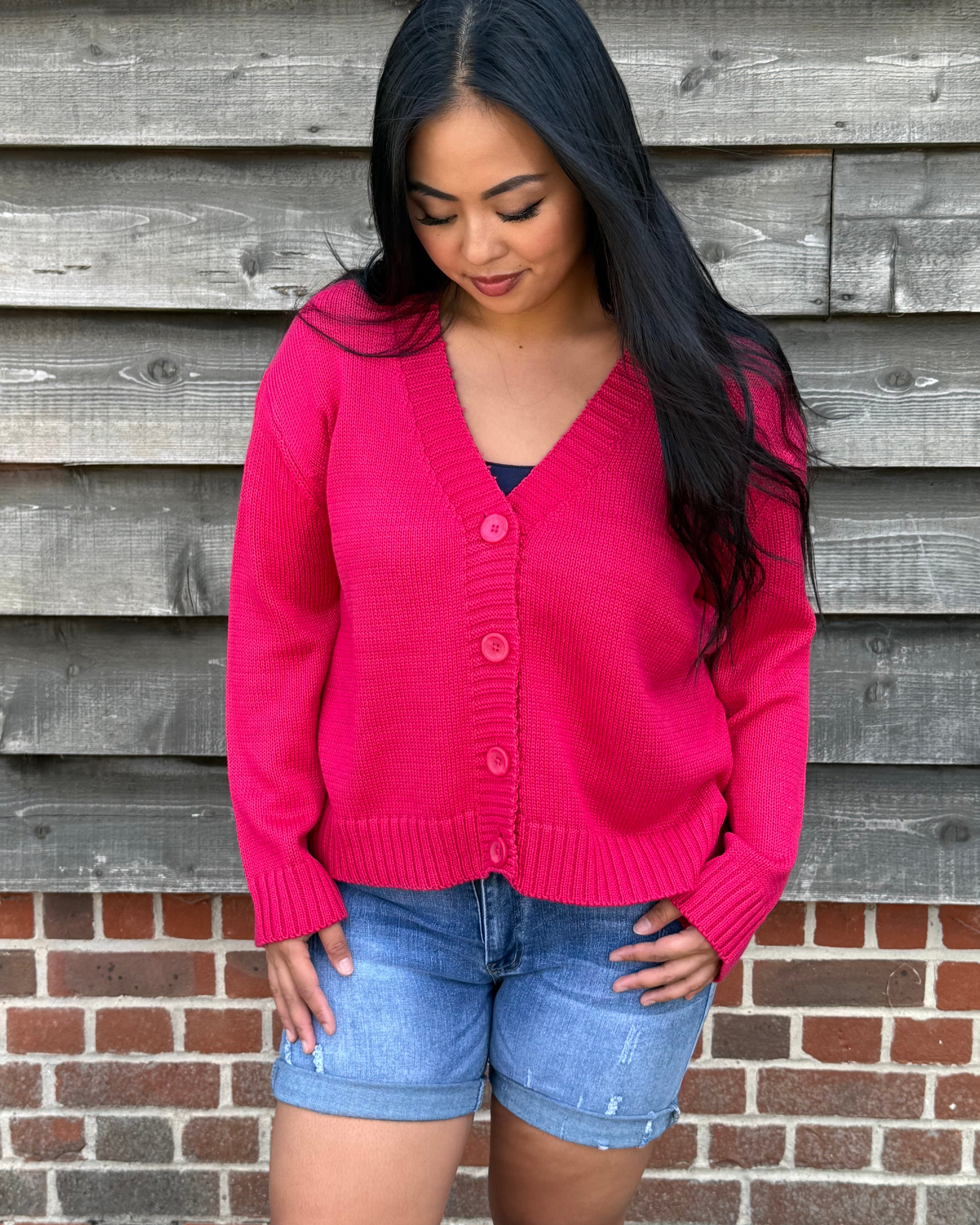 Woman wearing a pink cardigan and denim shorts standing against a wooden and brick wall.