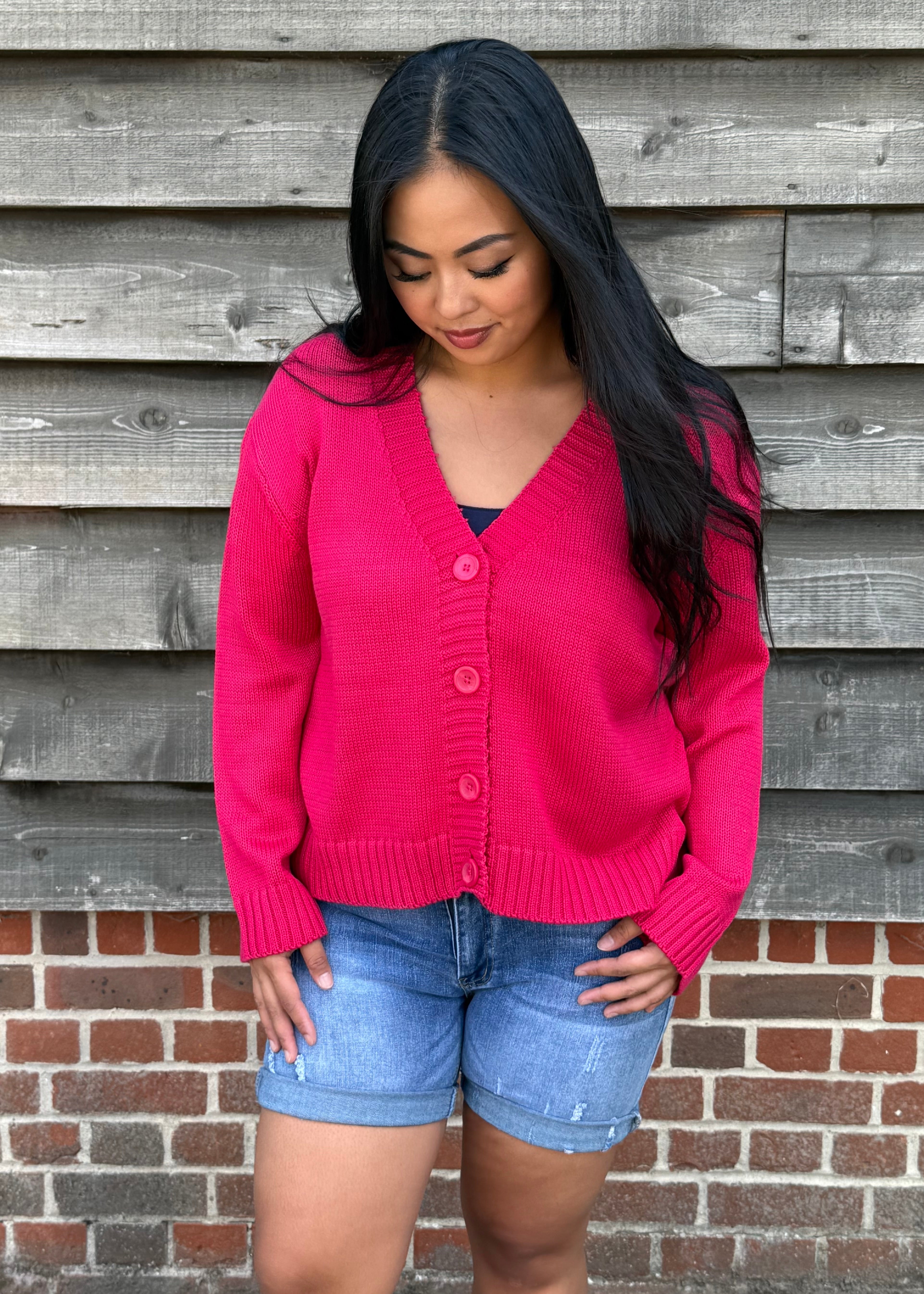 Woman wearing a pink cardigan and denim shorts standing against a wooden and brick wall.