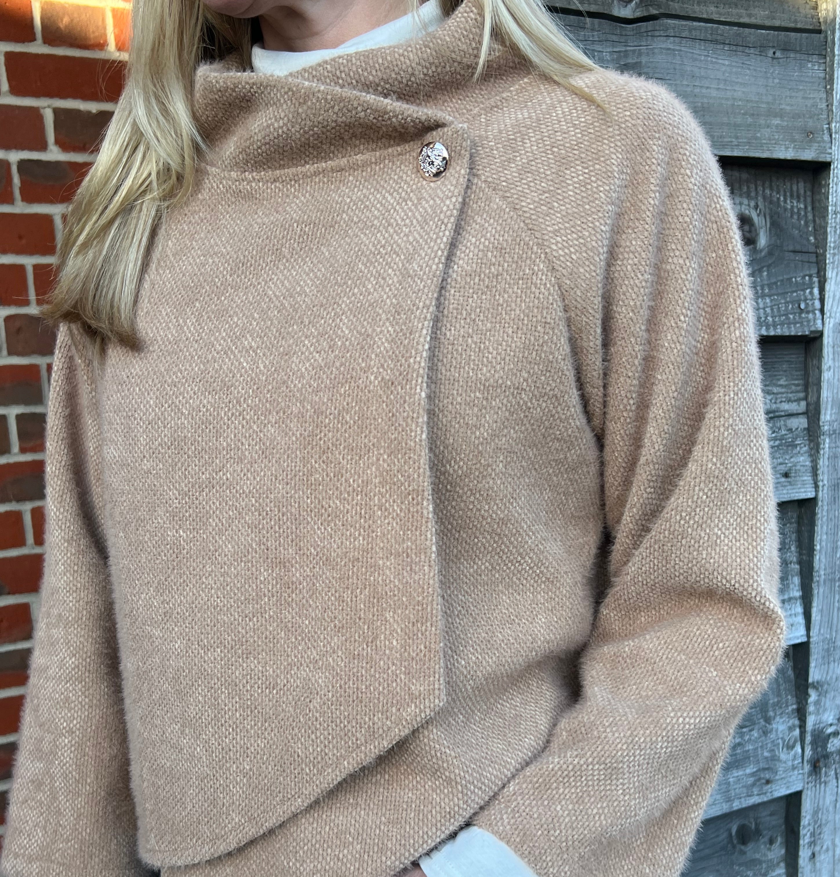 A woman standing outdoors wearing a beige knitted jacket and blue jeans.