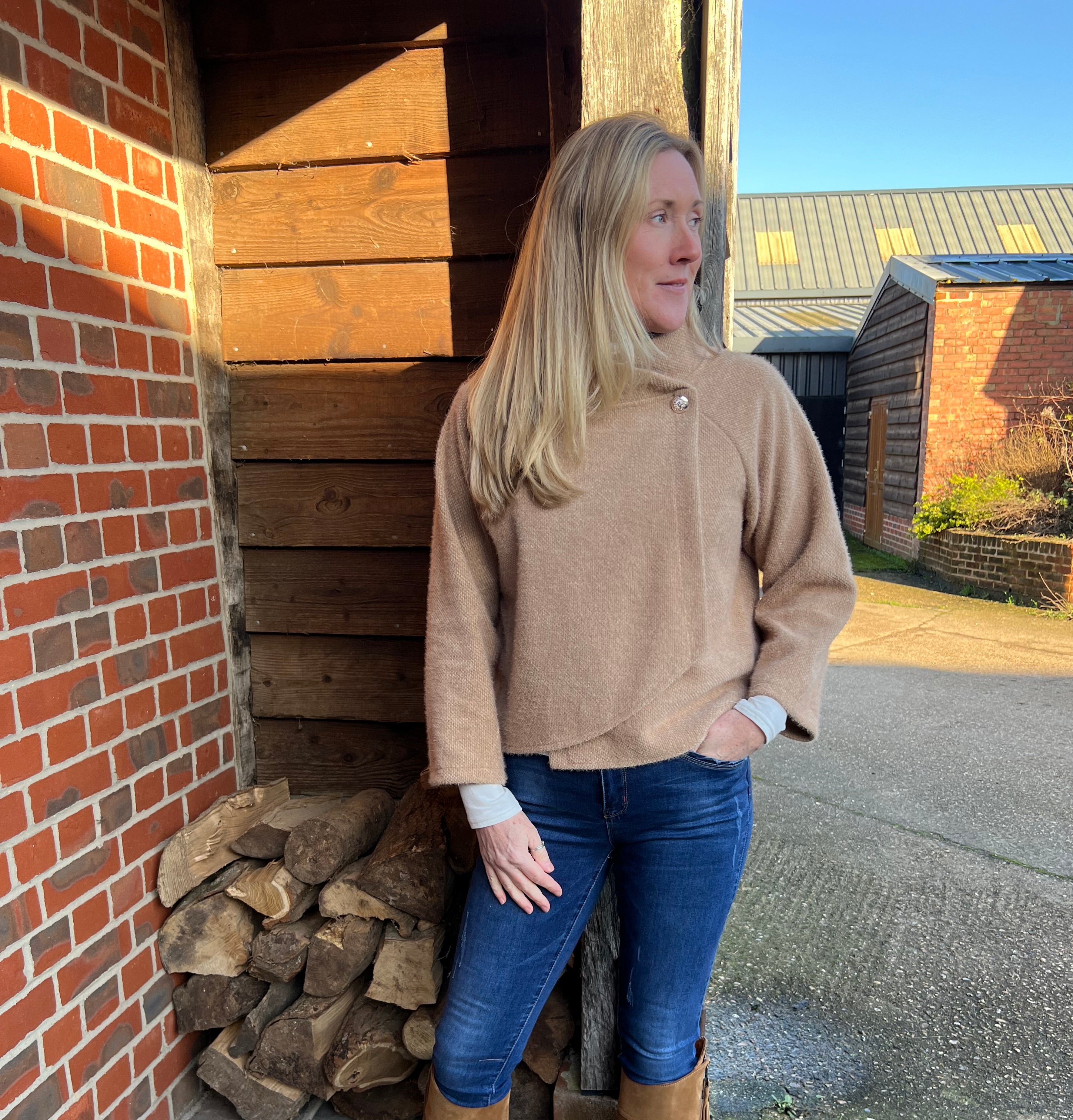 A woman standing outdoors wearing a beige knitted jacket and blue jeans, with brown boots.