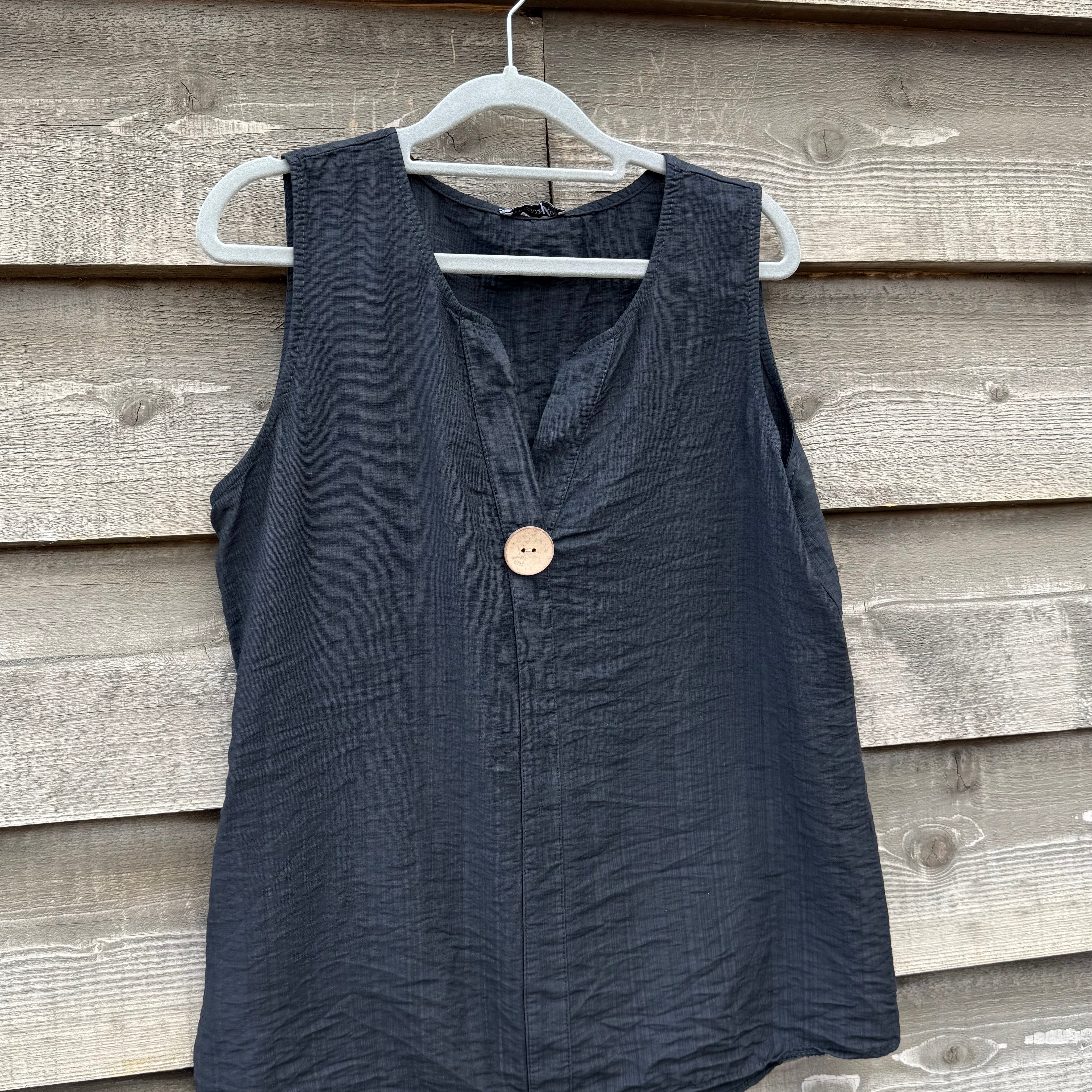 Black sleeveless top with a button on a hanger against a wooden wall.