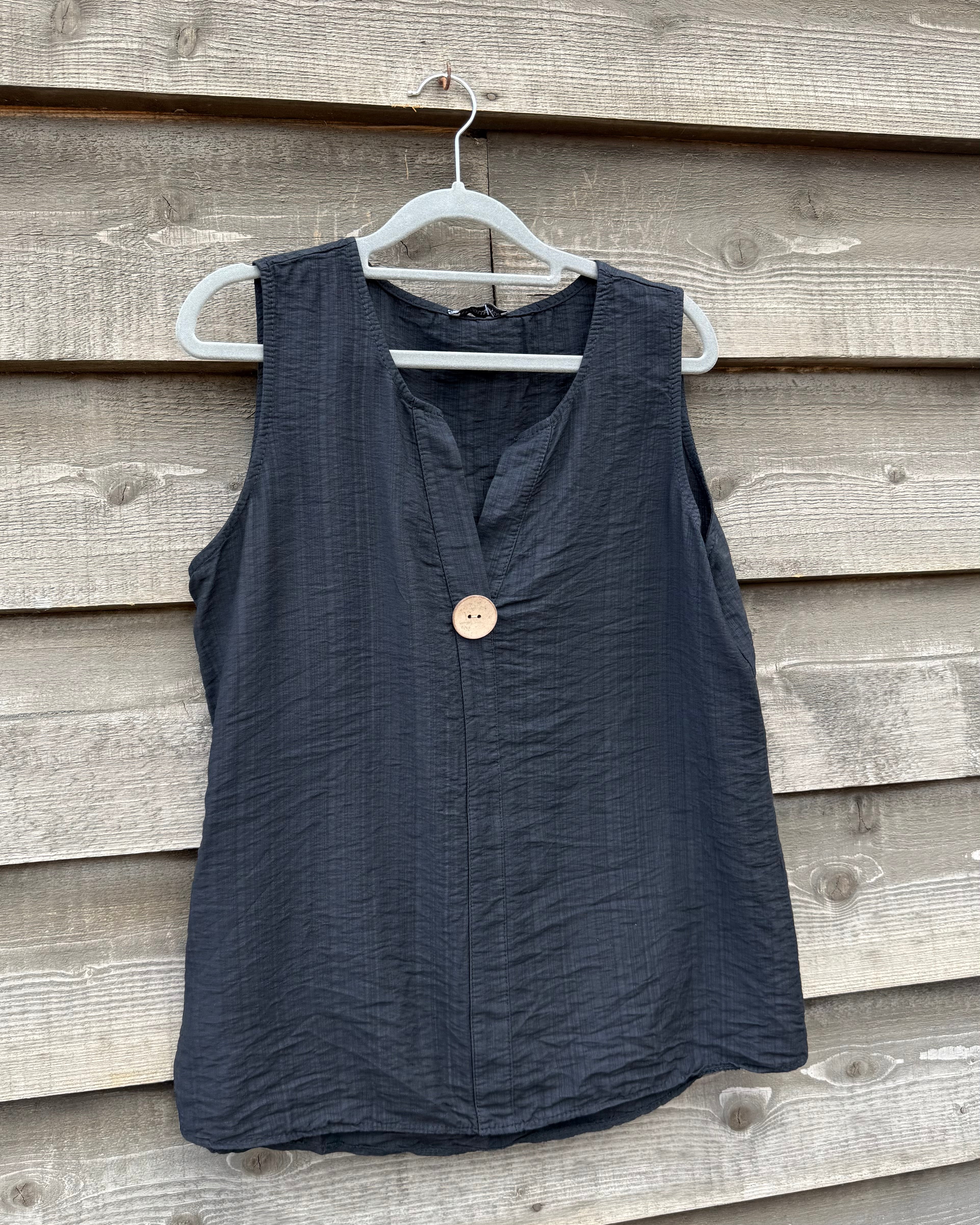 Black sleeveless top with a button on a hanger against a wooden wall.
