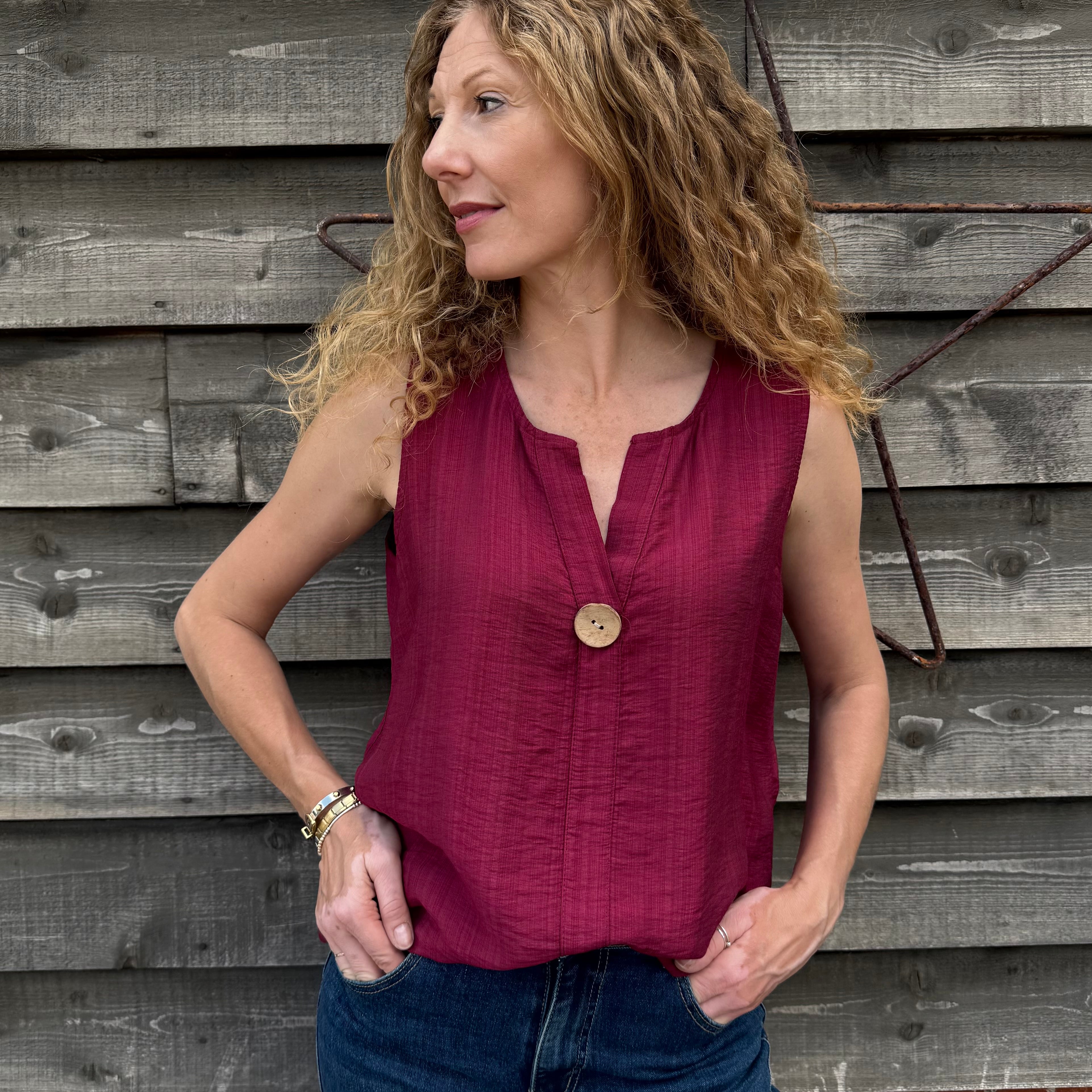 Woman wearing a red sleeveless top and blue jeans standing against a wooden wall.