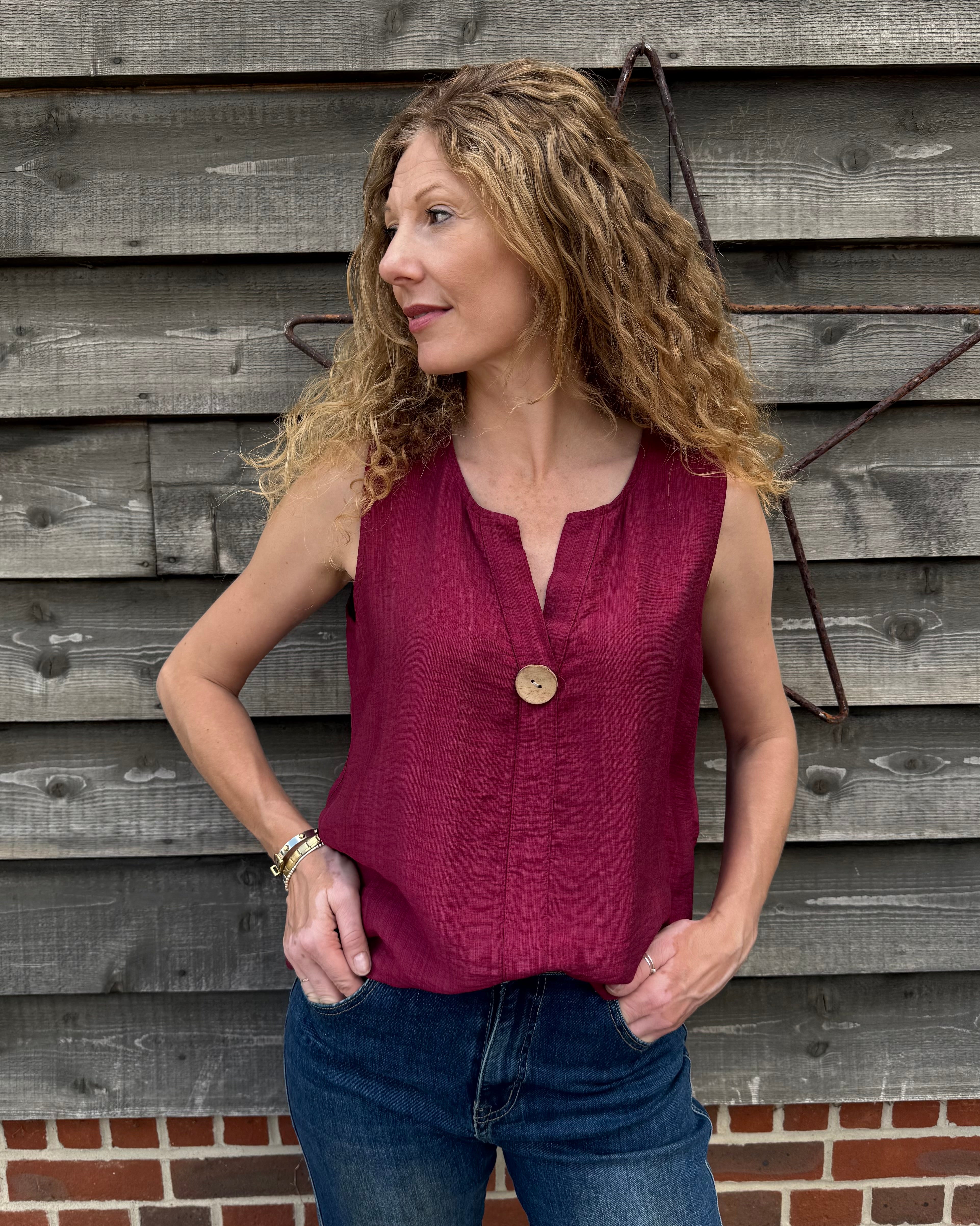 Woman wearing a red sleeveless top and blue jeans standing against a wooden wall.