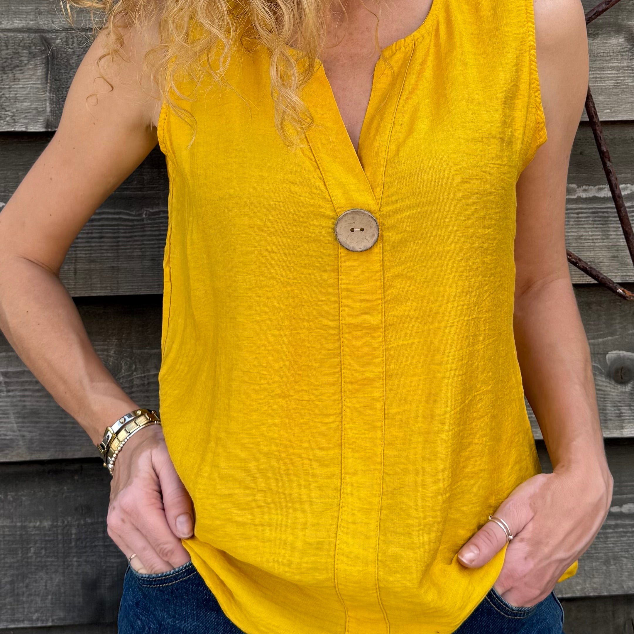 Woman wearing a yellow sleeveless top with a button, standing against a wooden background.