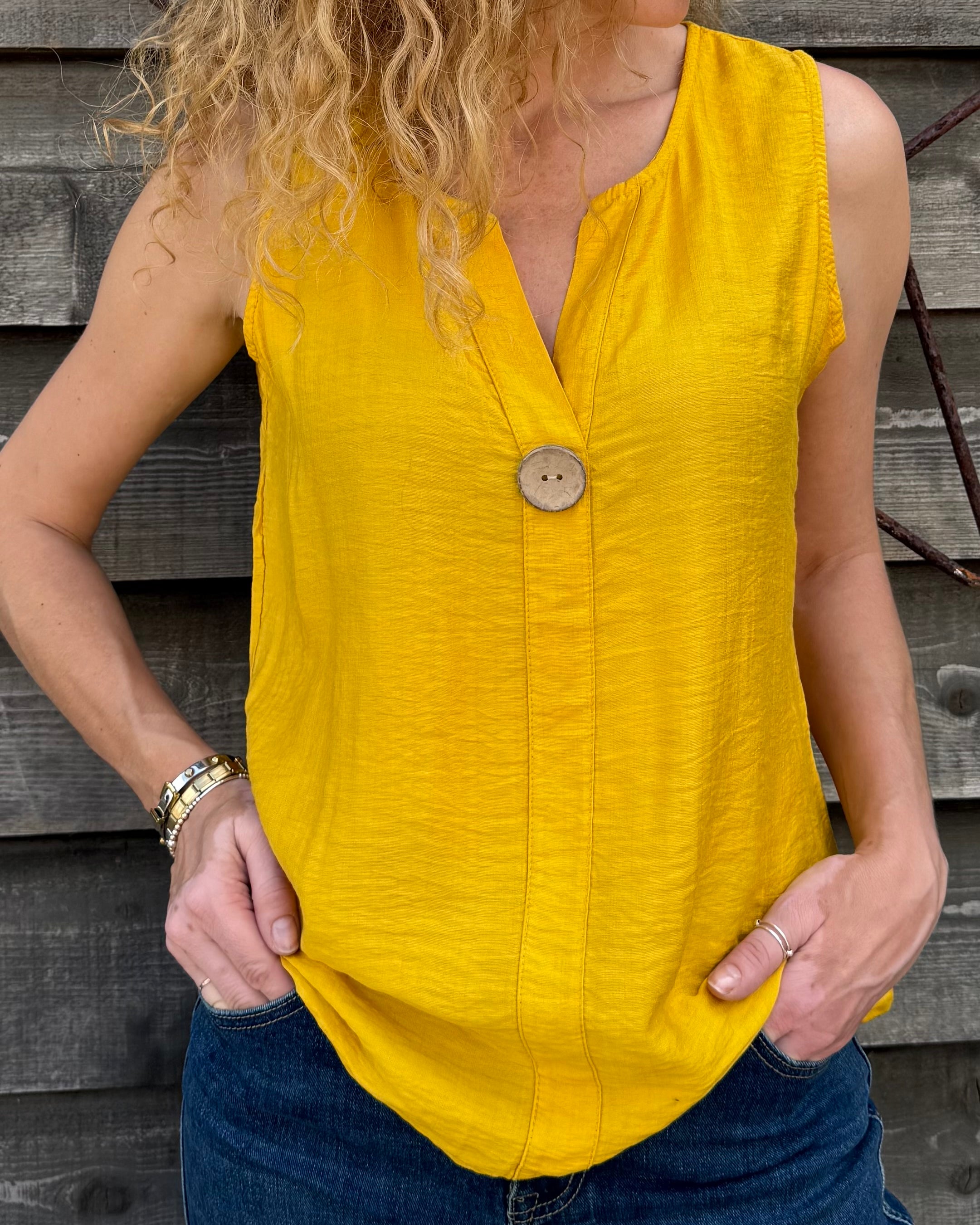 Woman wearing a yellow sleeveless top with a button, standing against a wooden background.