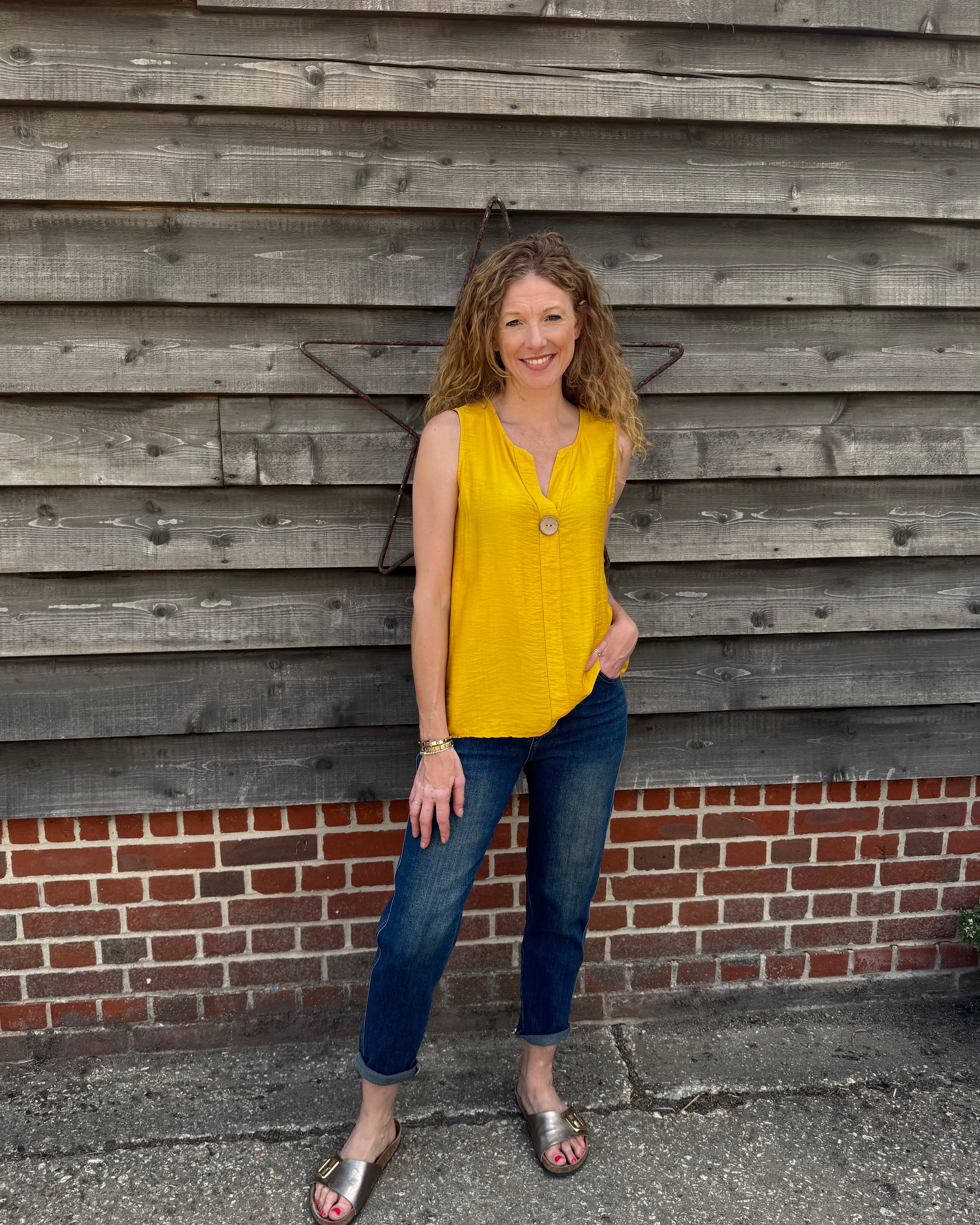 Woman in a yellow top and blue jeans standing against a wooden wall.