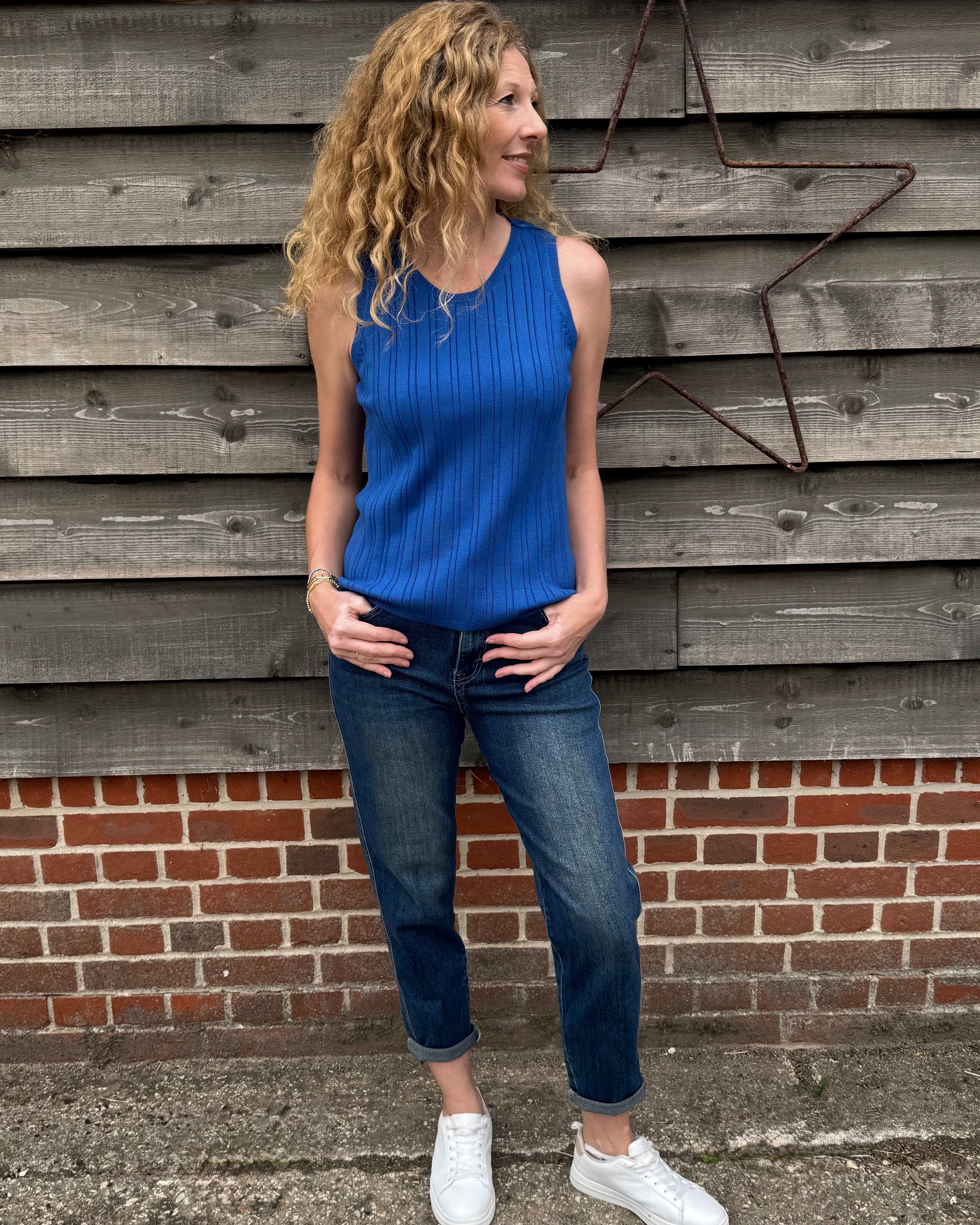 Woman wearing a blue sleeveless top and jeans standing against a wooden and brick wall.