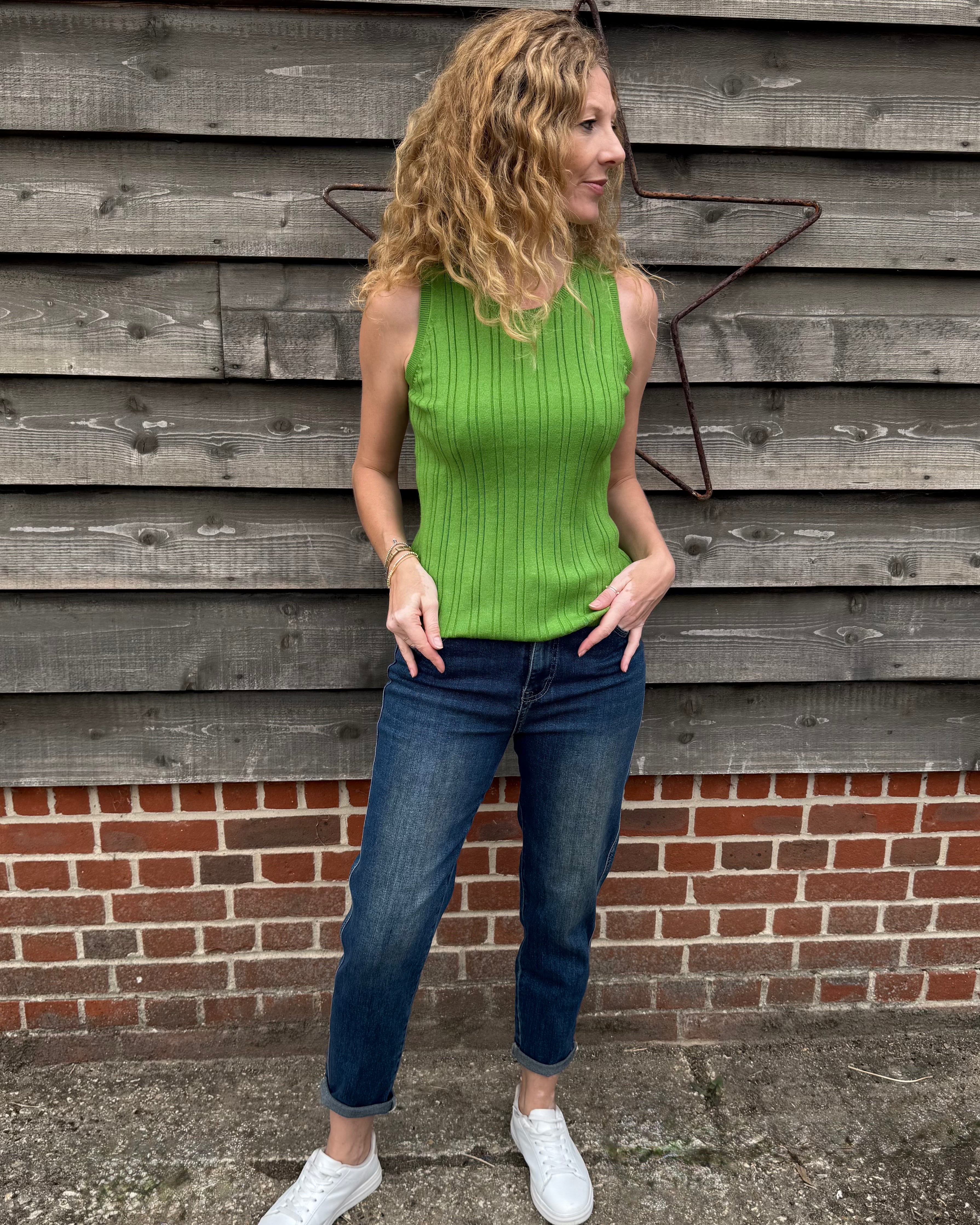 Woman wearing a green sleeveless top and blue jeans standing against a wooden wall.