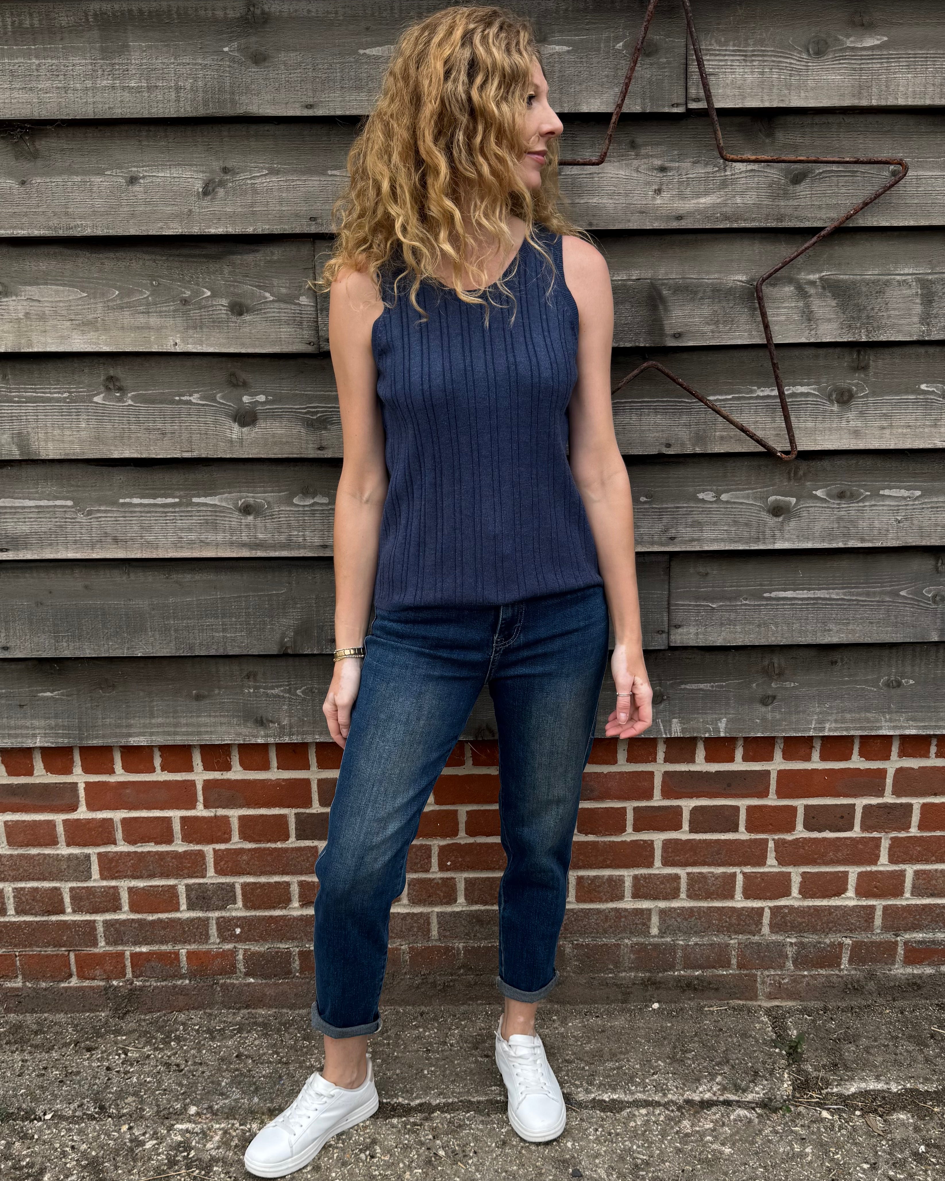 Woman wearing a blue sleeveless top and jeans standing against a wooden wall.