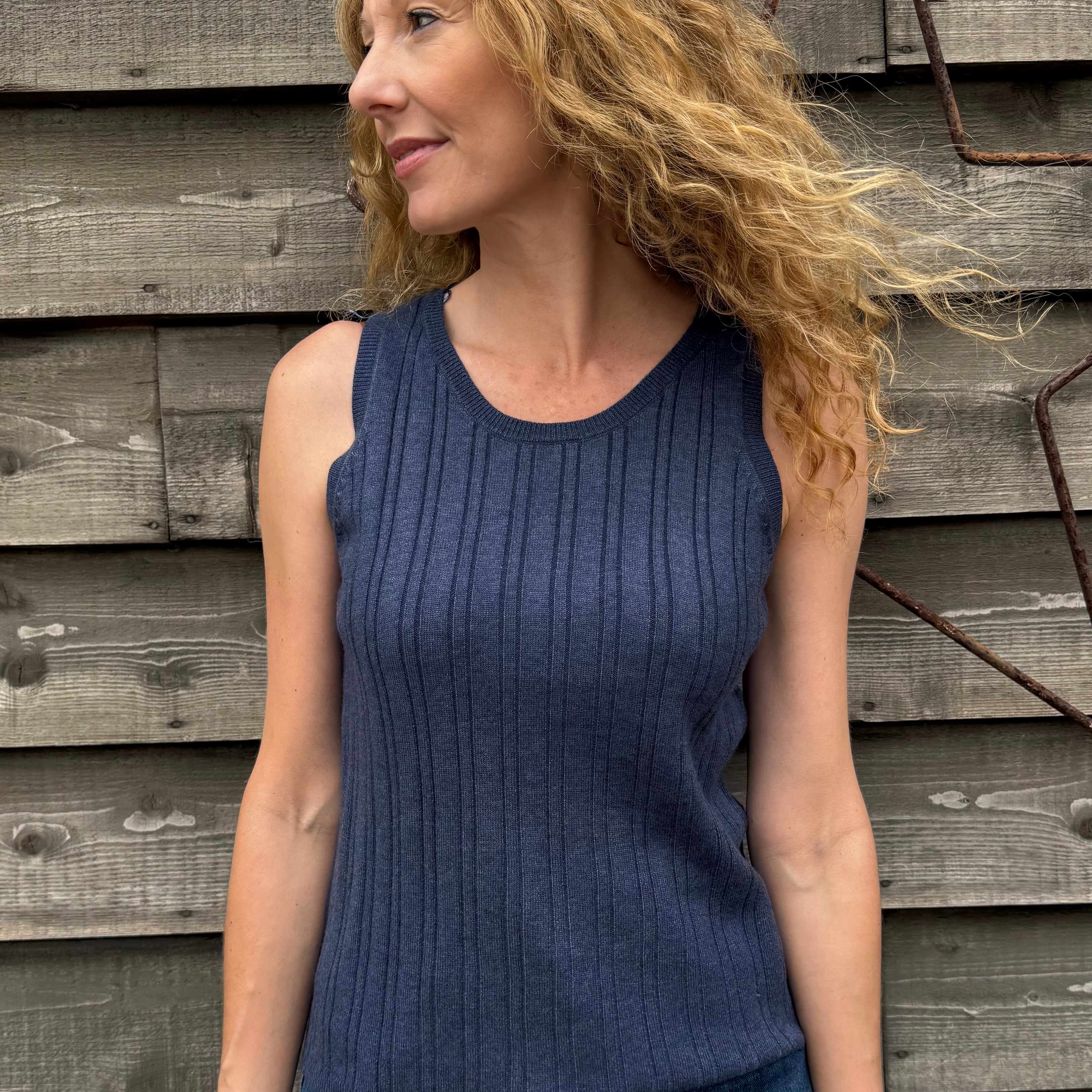 Woman wearing a blue sleeveless top and jeans standing against a wooden wall.