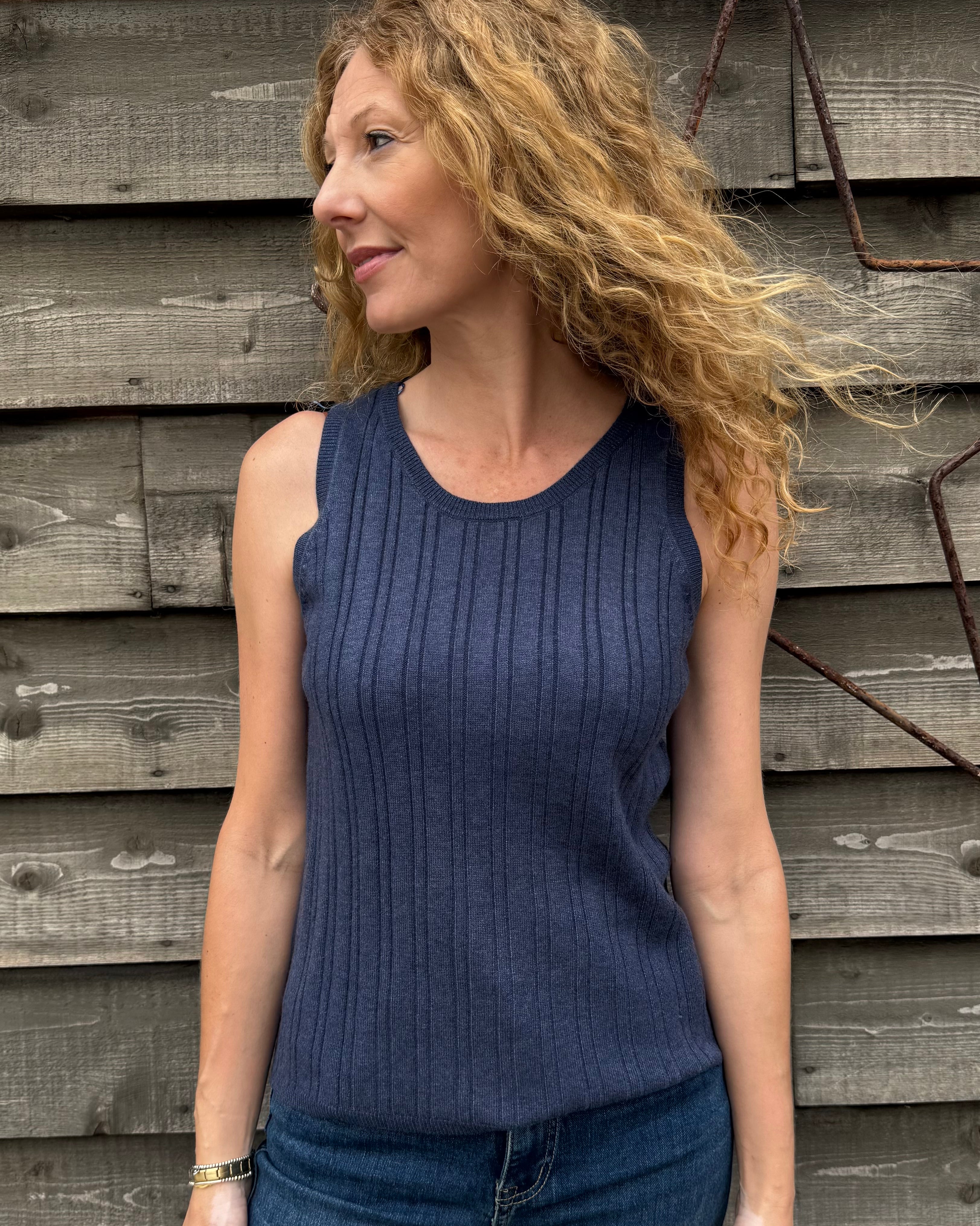 Woman wearing a blue sleeveless top and jeans standing against a wooden wall.