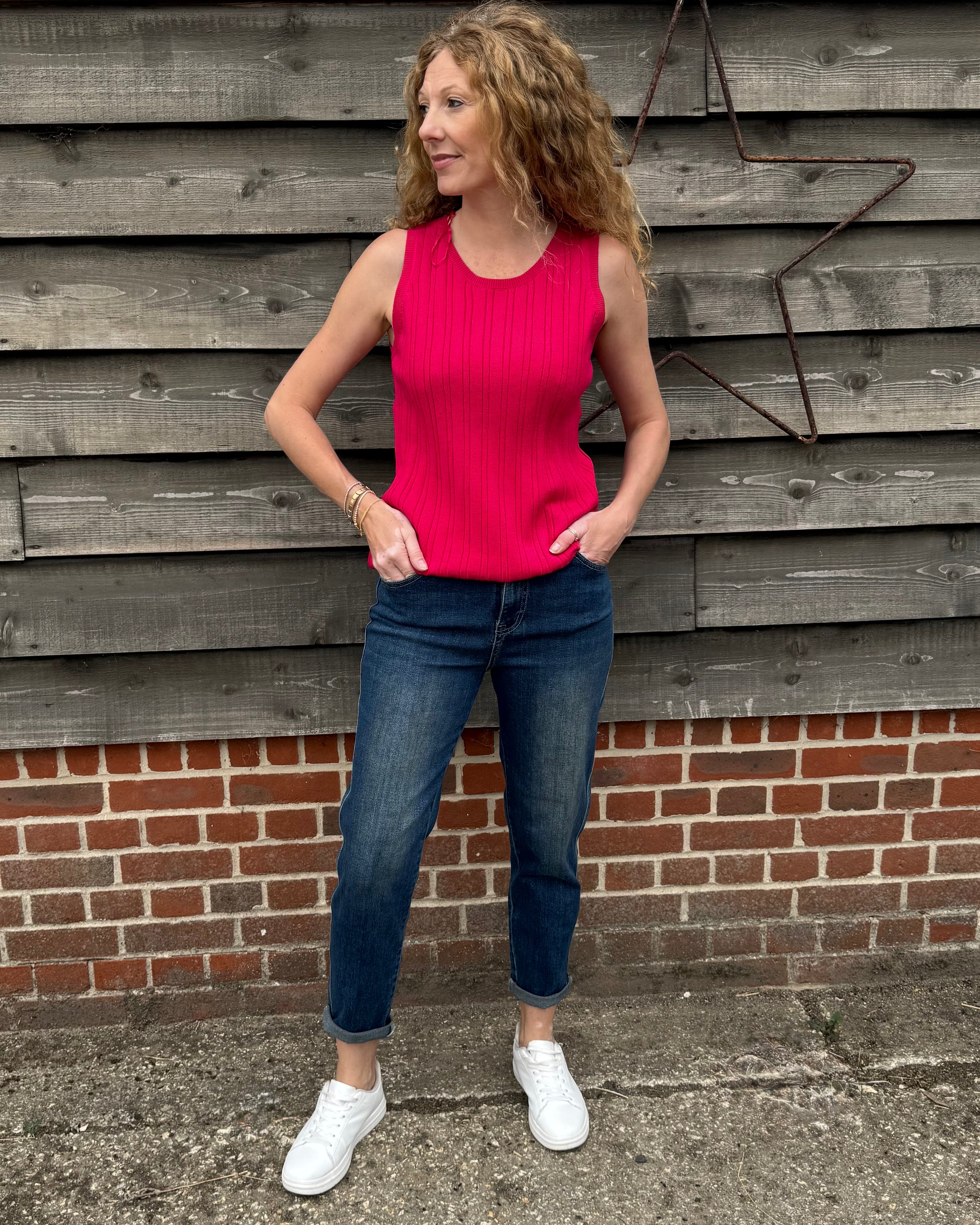 Woman wearing a pink top and blue jeans standing against a wooden wall.