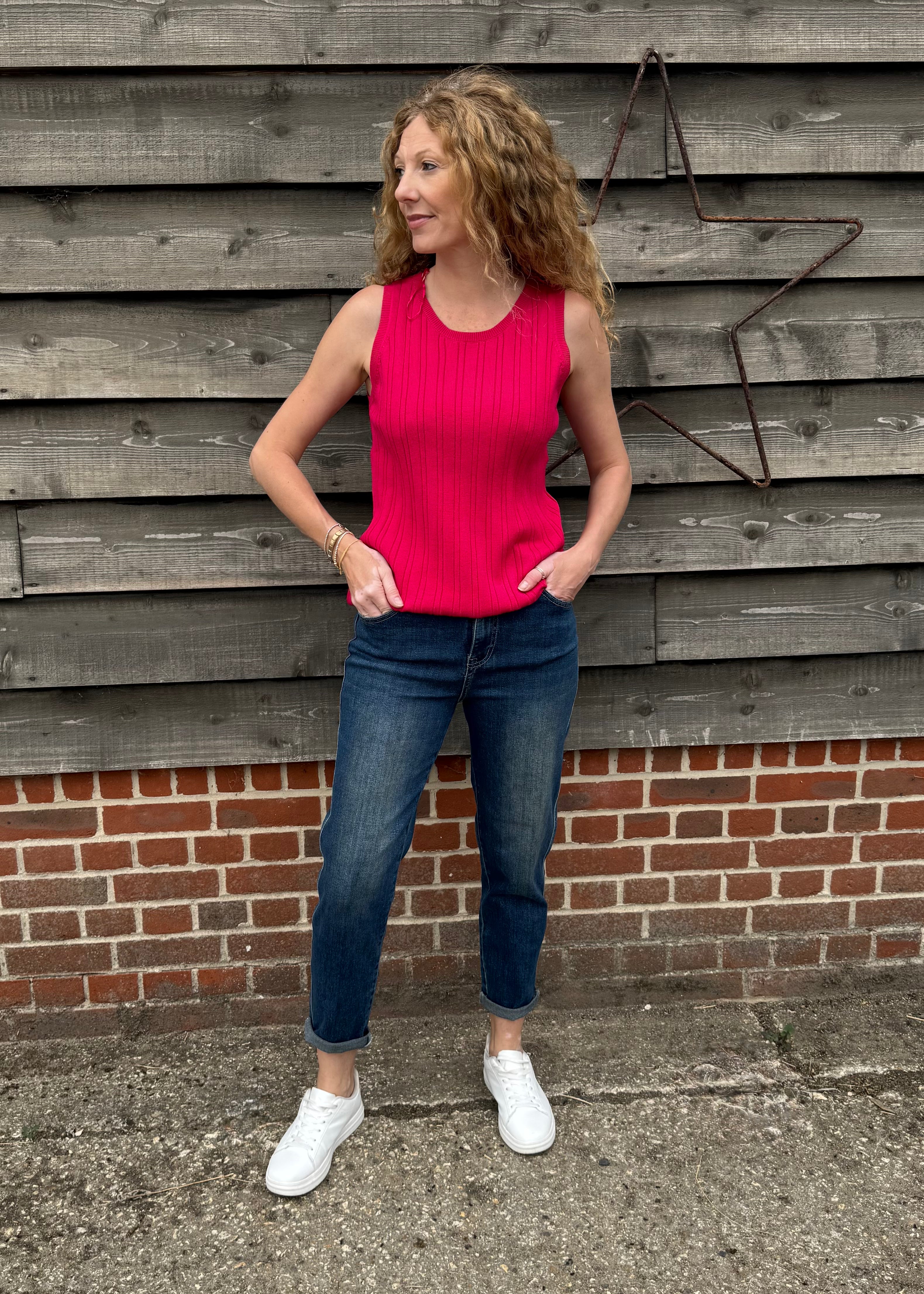 Woman wearing a pink top and blue jeans standing against a wooden wall.