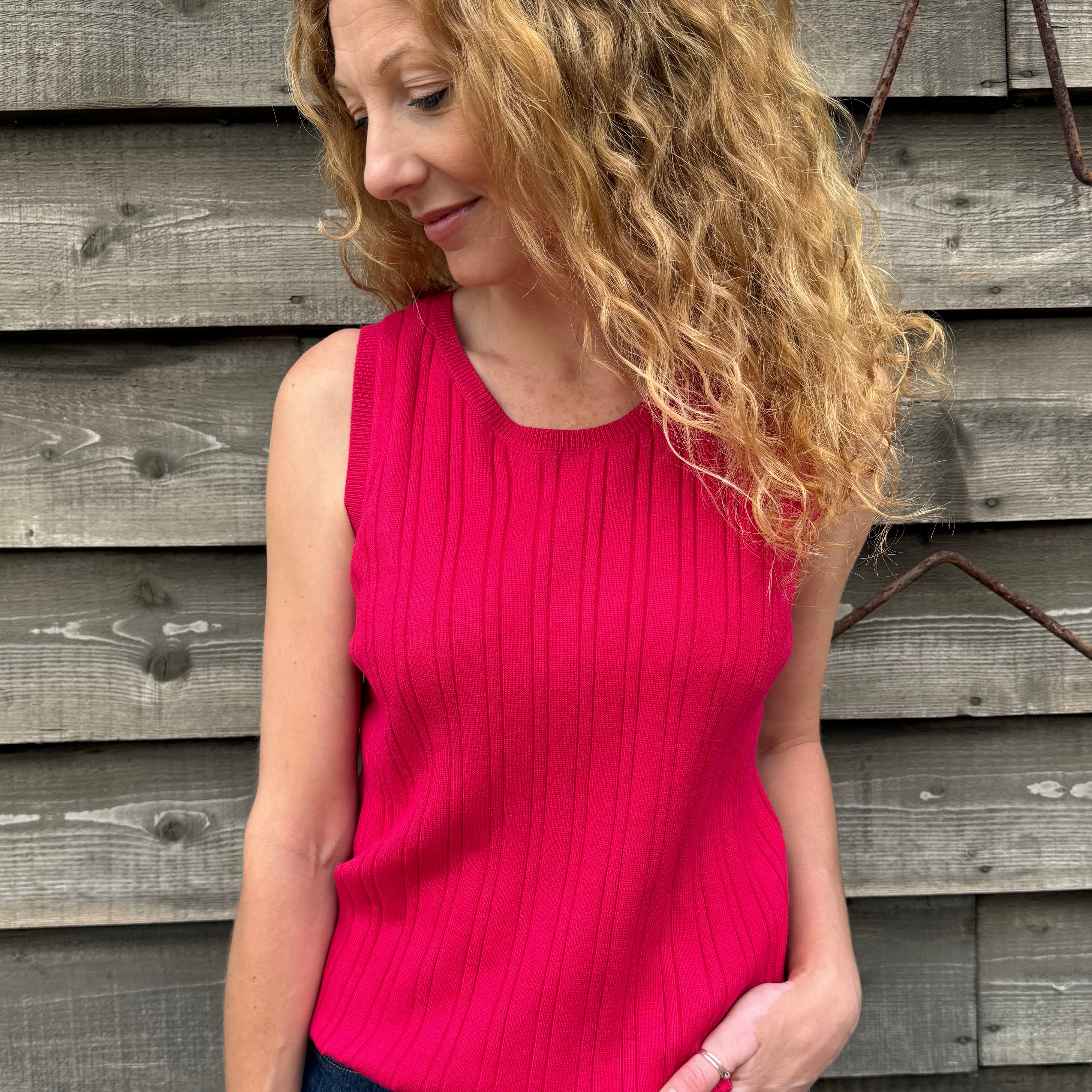 Woman wearing a pink sleeveless top and blue jeans standing against a wooden wall.