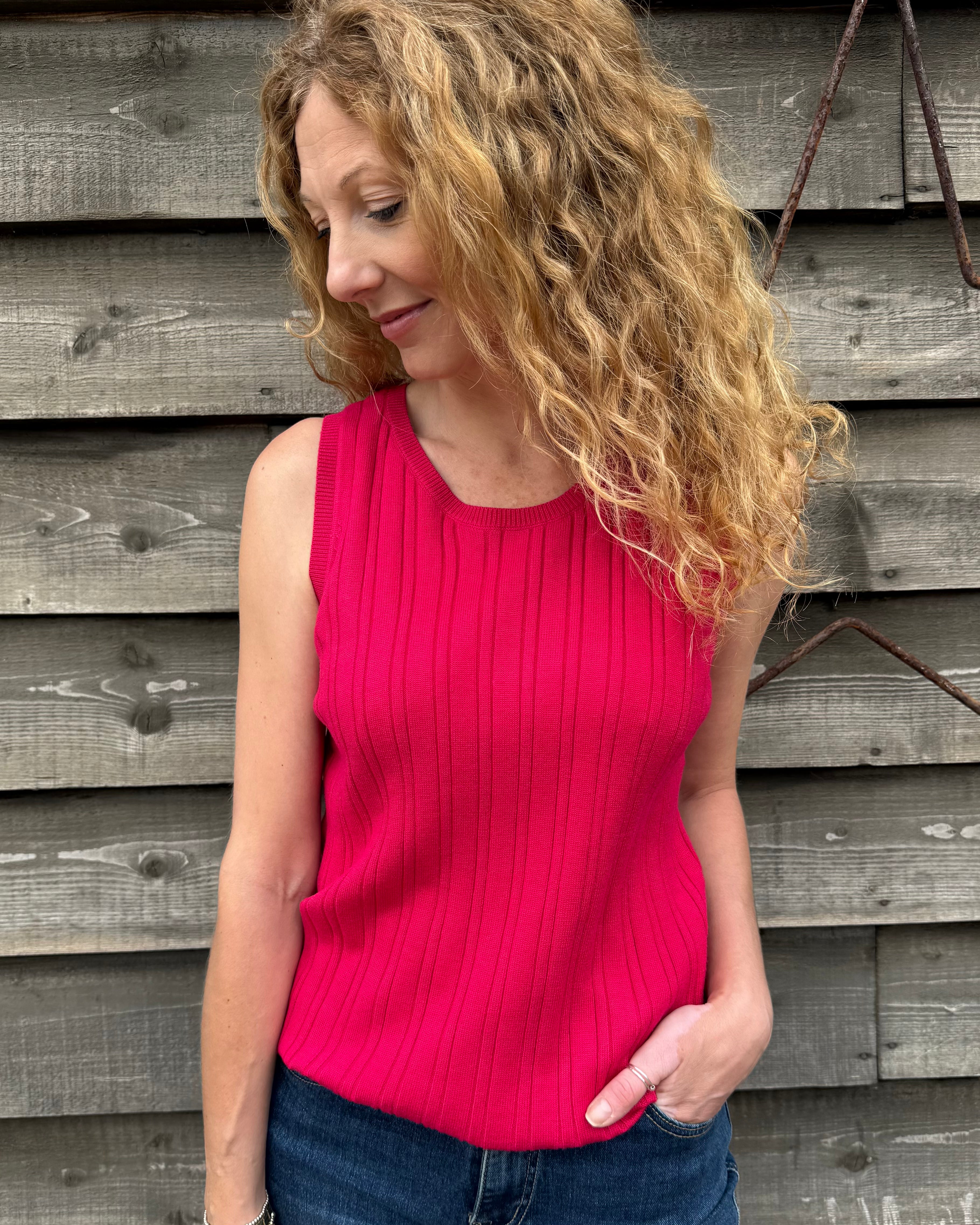 Woman wearing a pink sleeveless top and blue jeans standing against a wooden wall.