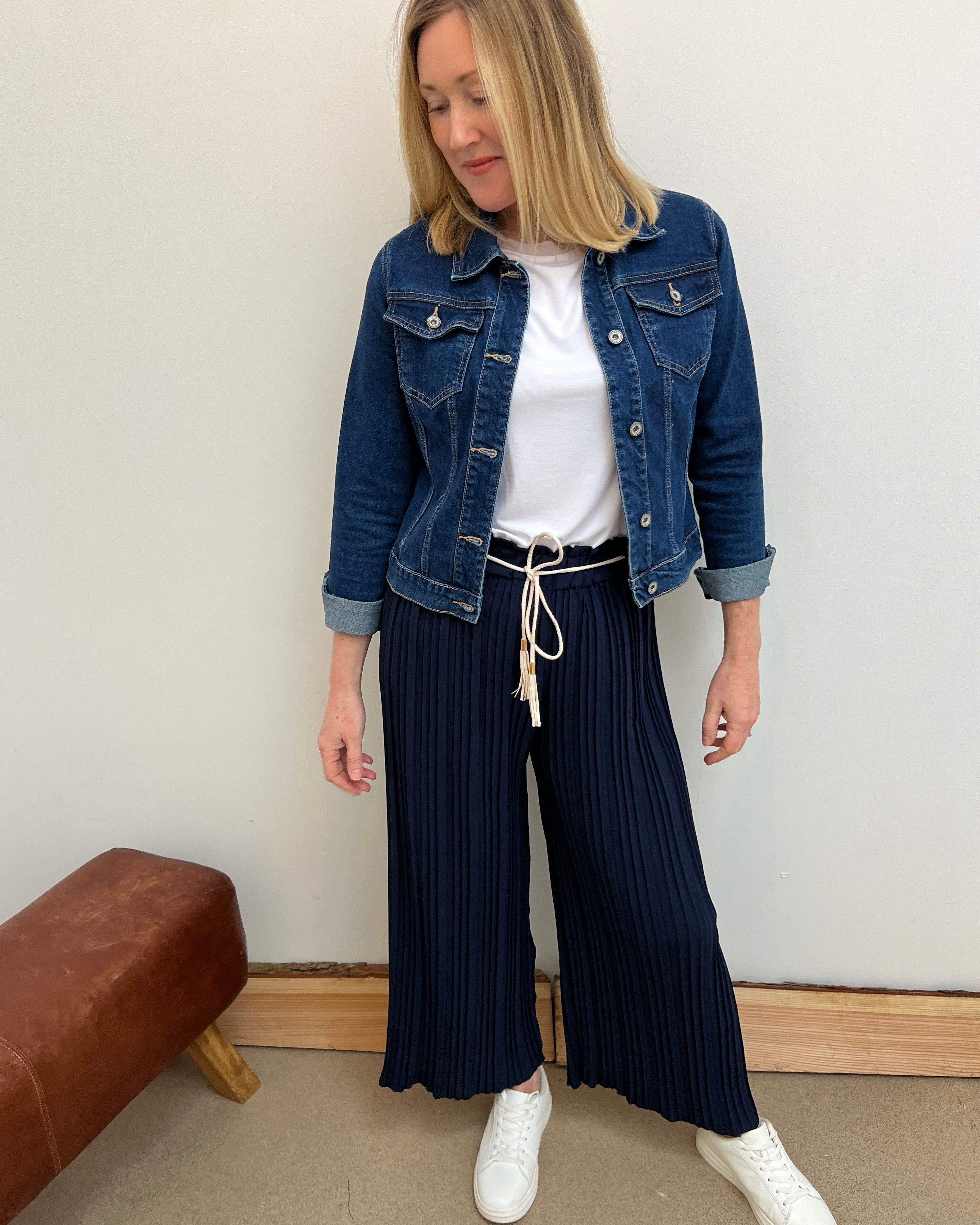 Woman wearing a denim jacket, white shirt, and navy pleated pants against a plain wall.