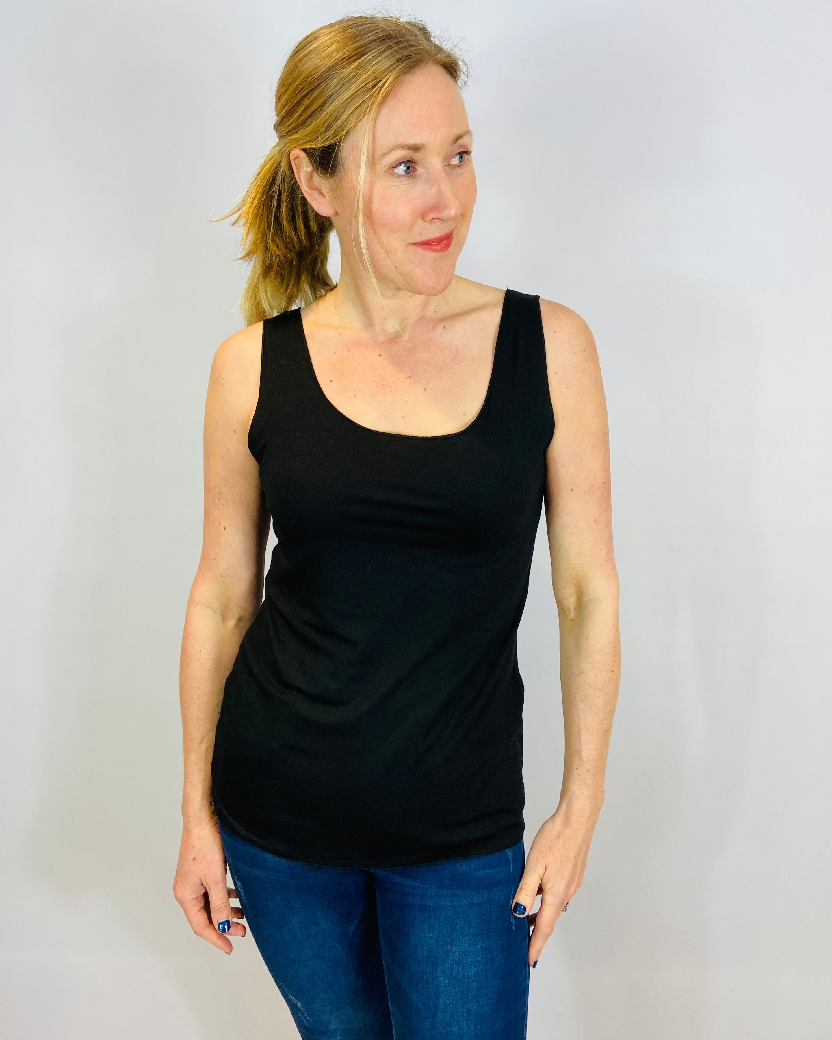 A woman wearing a black sleeveless tank top and blue jeans standing against a plain background.