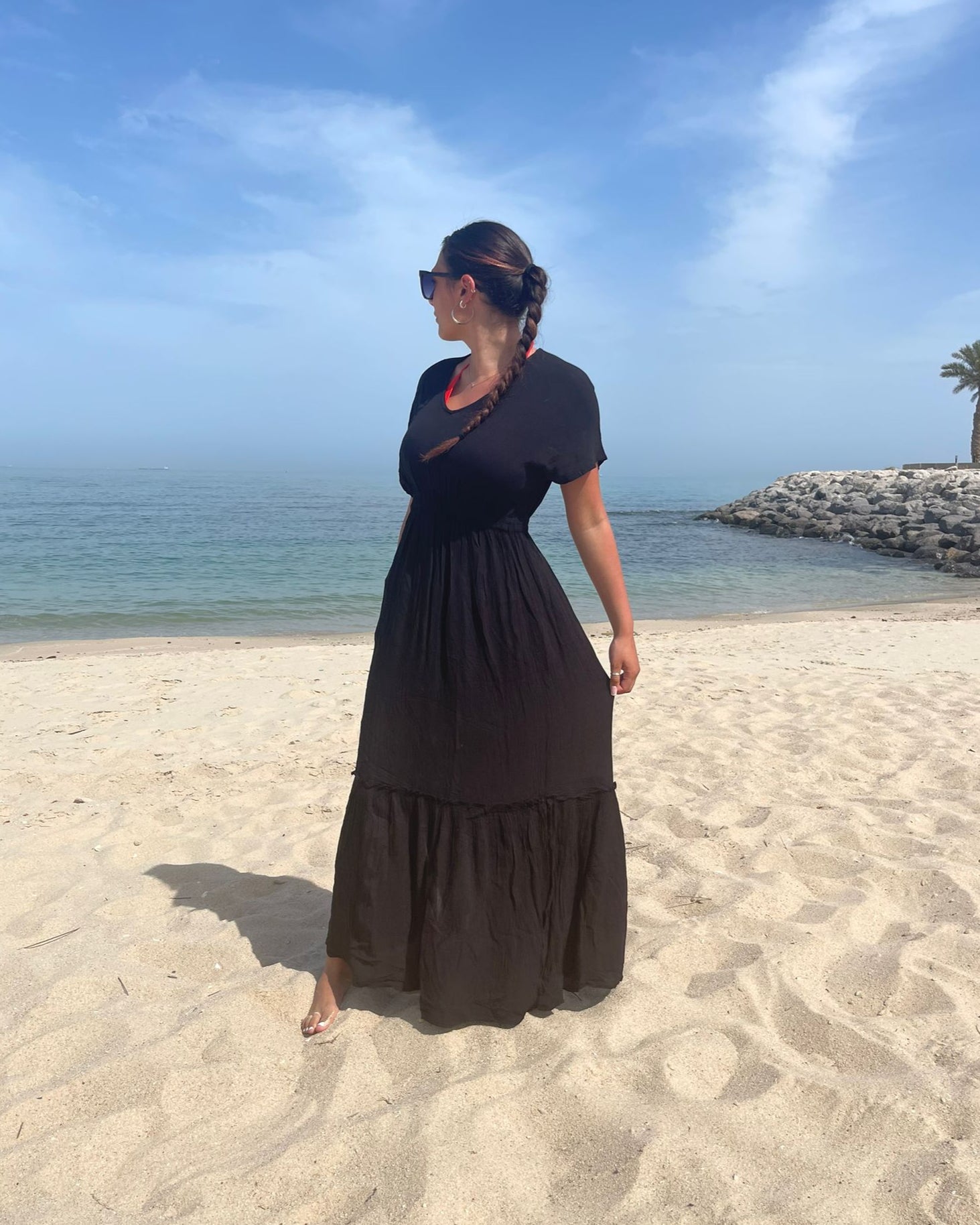 A person standing on a sandy beach wearing a navy maxi dress with short sleeves and a knee-length skirt, with a clear blue sky and some palm trees in the background.