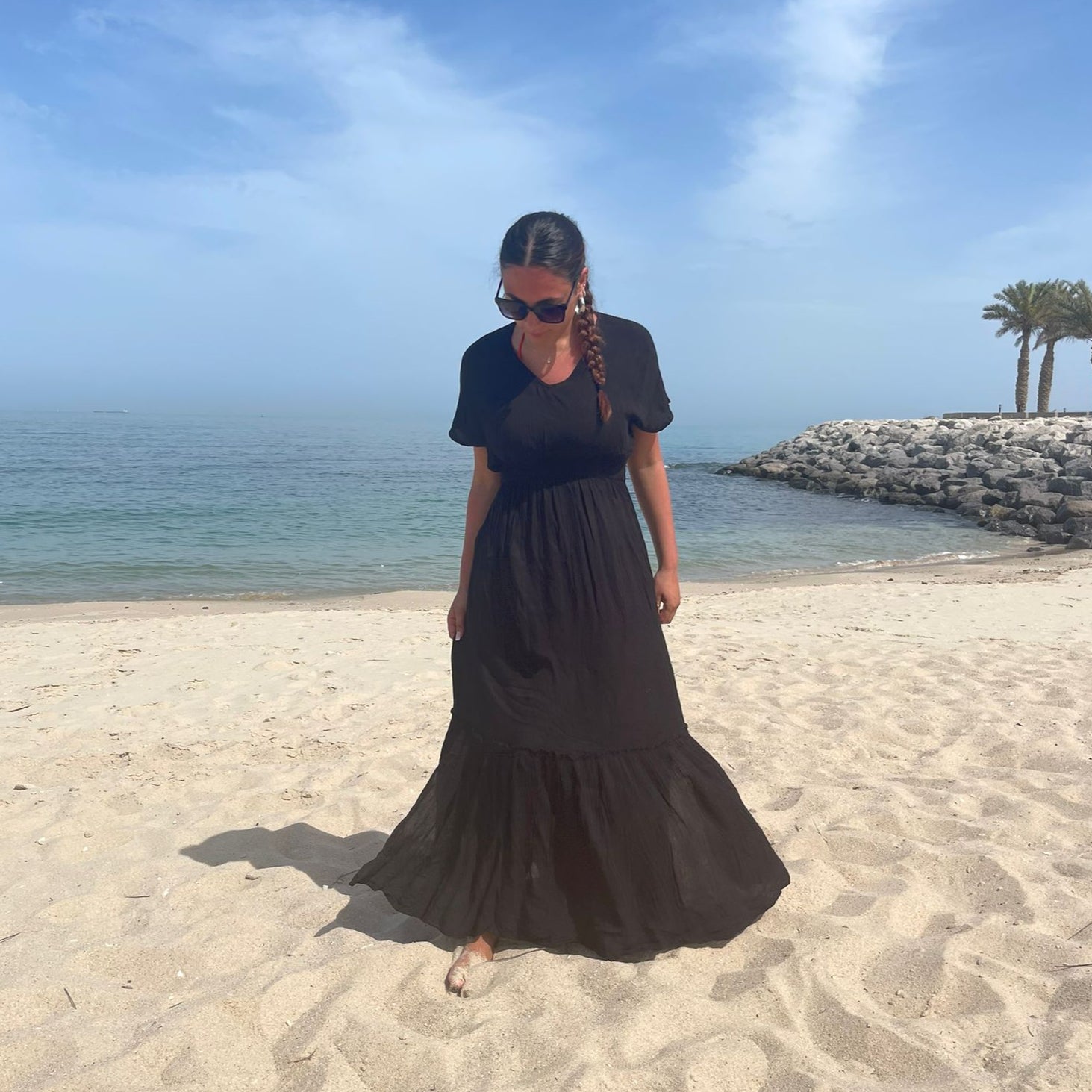 A person standing on a sandy beach wearing a navy maxi dress with short sleeves and a knee-length skirt, with a clear blue sky and some palm trees in the background.