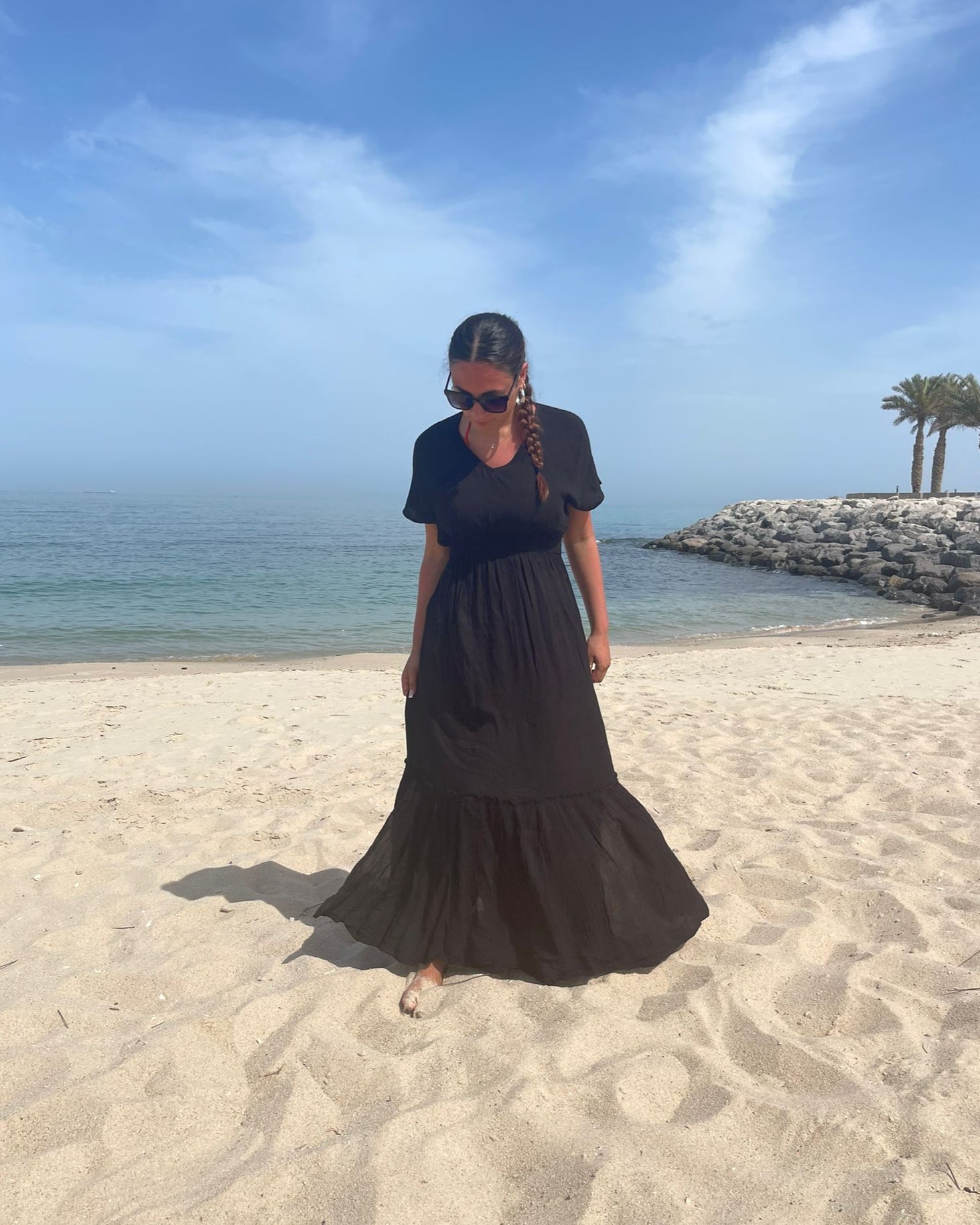 A person standing on a sandy beach wearing a navy maxi dress with short sleeves and a knee-length skirt, with a clear blue sky and some palm trees in the background.