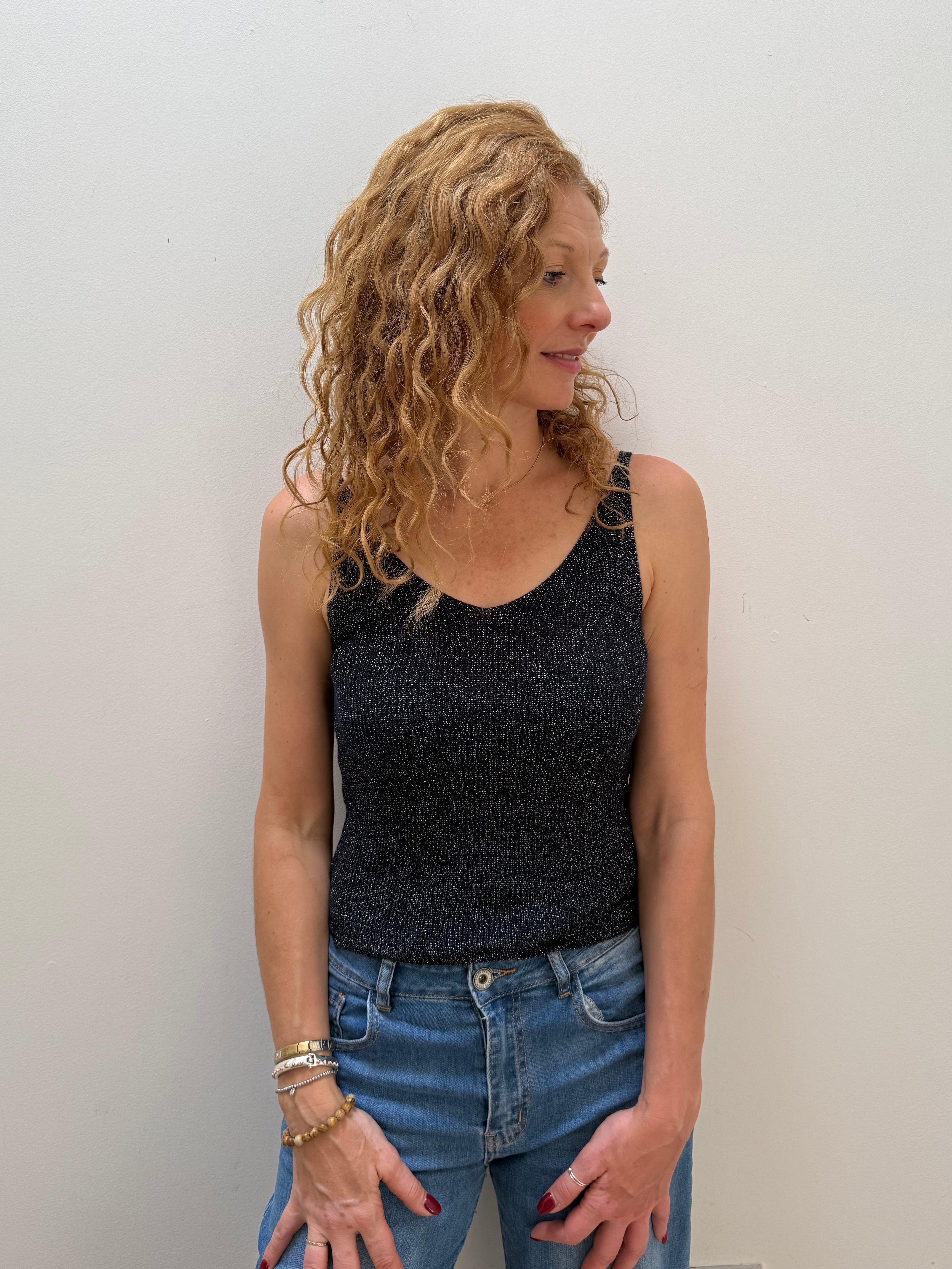Woman with curly hair wearing a black sleeveless top and blue jeans against a white wall.
