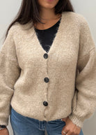 A woman wearing a beige, textured knit cardigan with dark buttons over a black top and blue jeans, standing against a plain light gray wall.