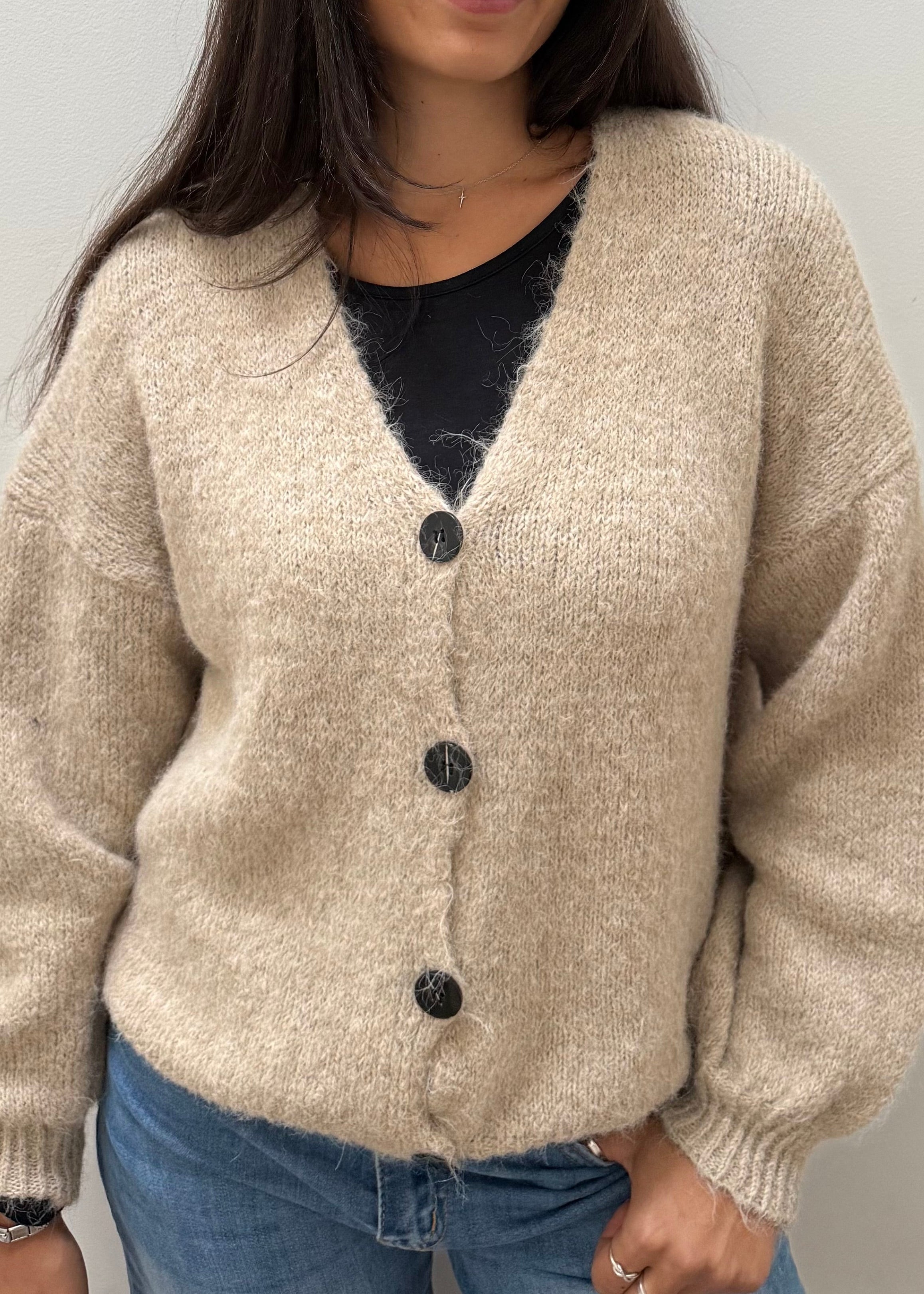A woman wearing a beige, textured knit cardigan with dark buttons over a black top and blue jeans, standing against a plain light gray wall.