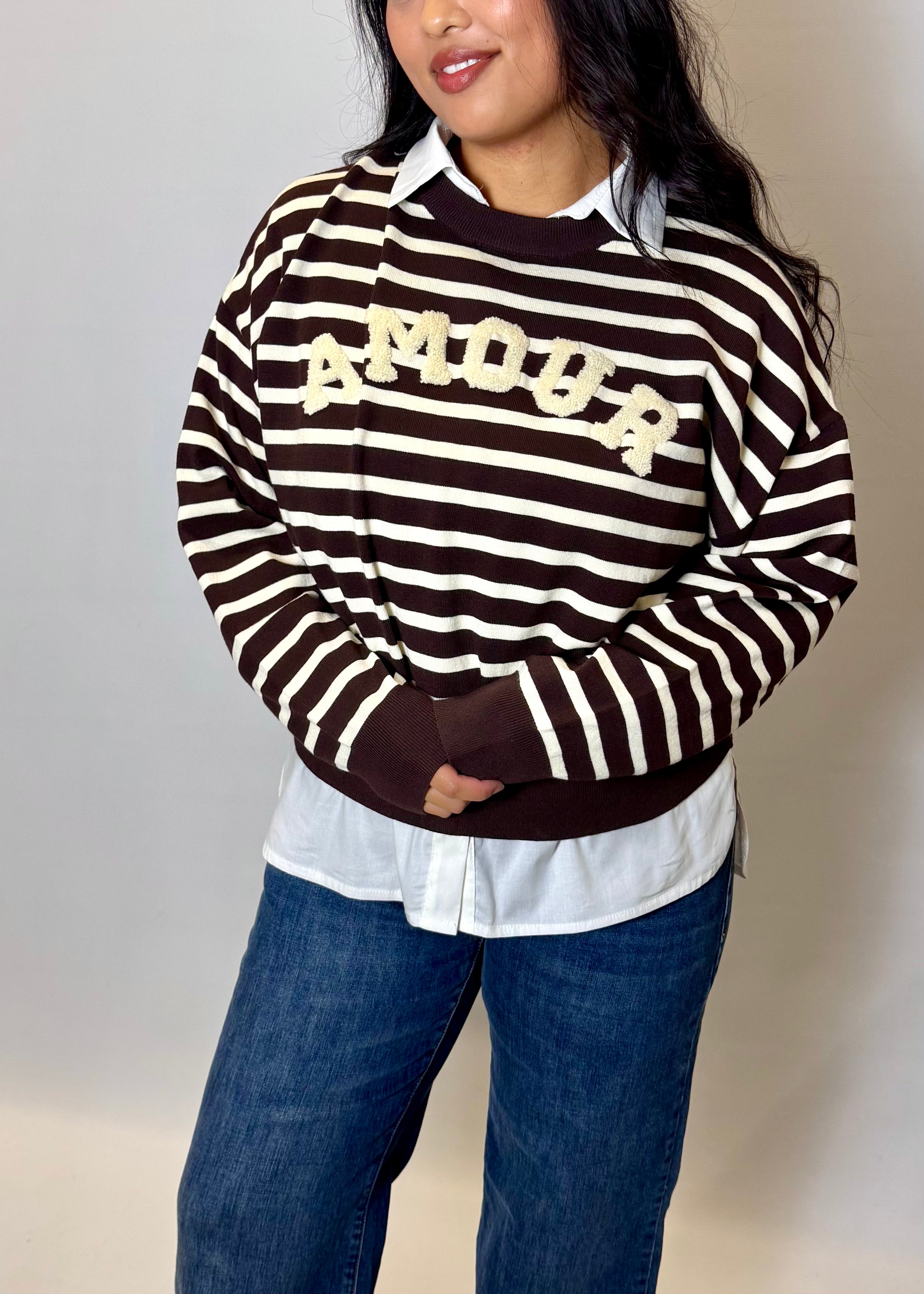Person wearing a black and white striped sweater with 'MOOP' text, white shirt, and blue jeans against a plain background.