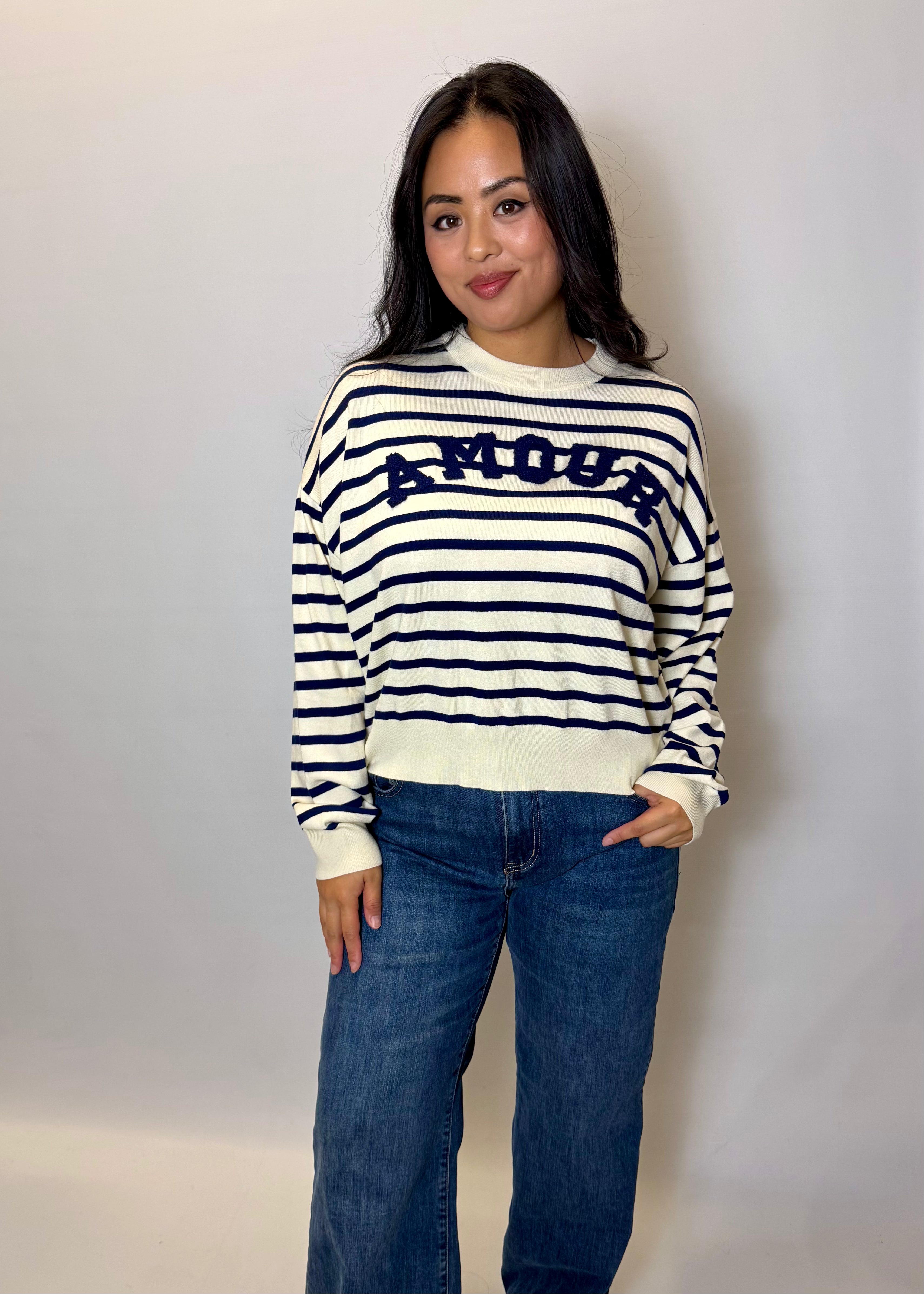 Woman wearing a striped sweater with 'AMOUR' text and jeans against a plain background