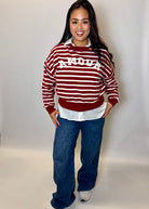 Woman wearing a red and white striped sweater with 'AMOUR' text, white shirt, blue jeans, and white shoes on a plain background.