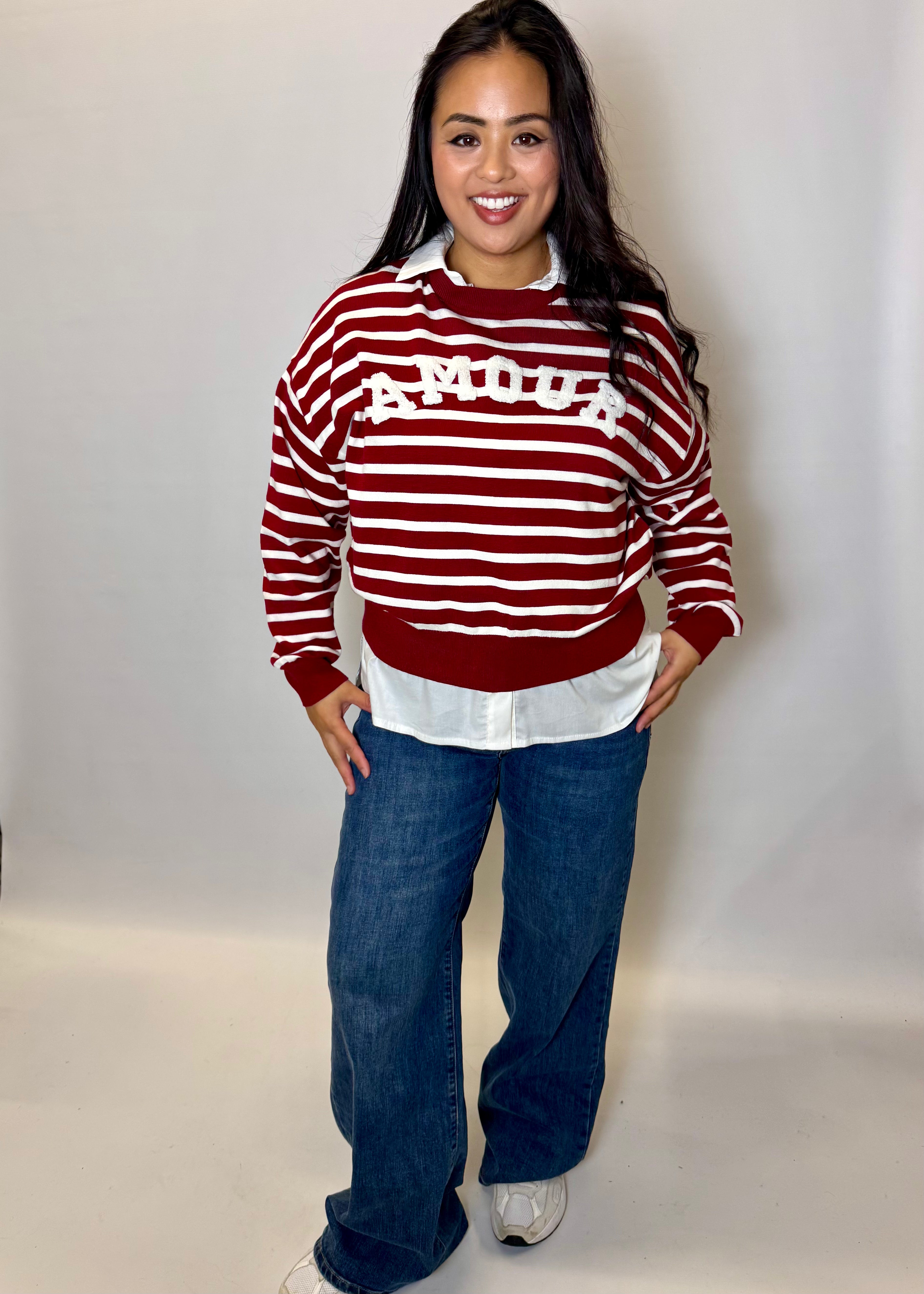 Woman wearing a red and white striped sweater with 'AMOUR' text, white shirt, blue jeans, and white shoes on a plain background.