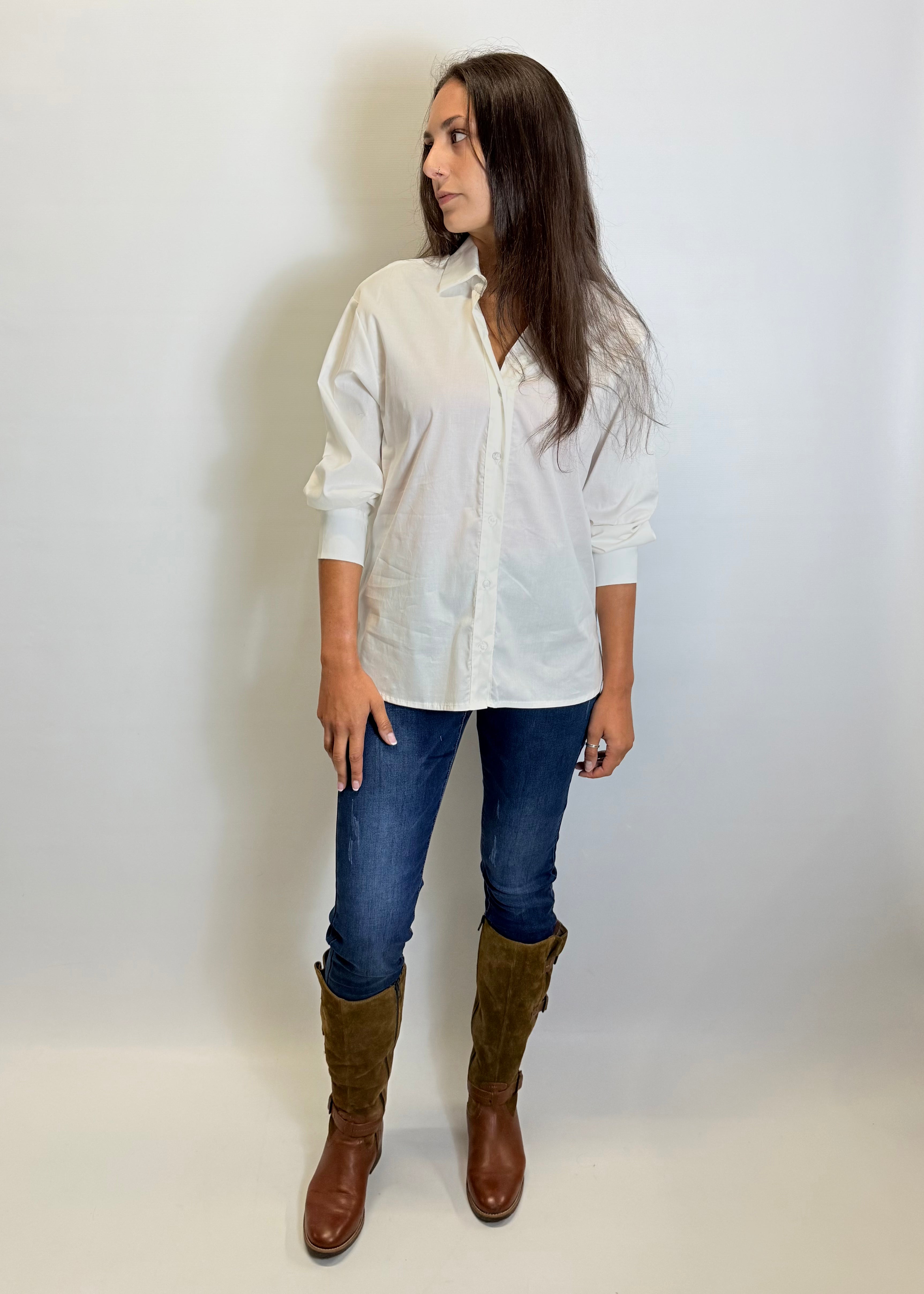 Woman wearing a white shirt, blue jeans, and brown boots against a plain background