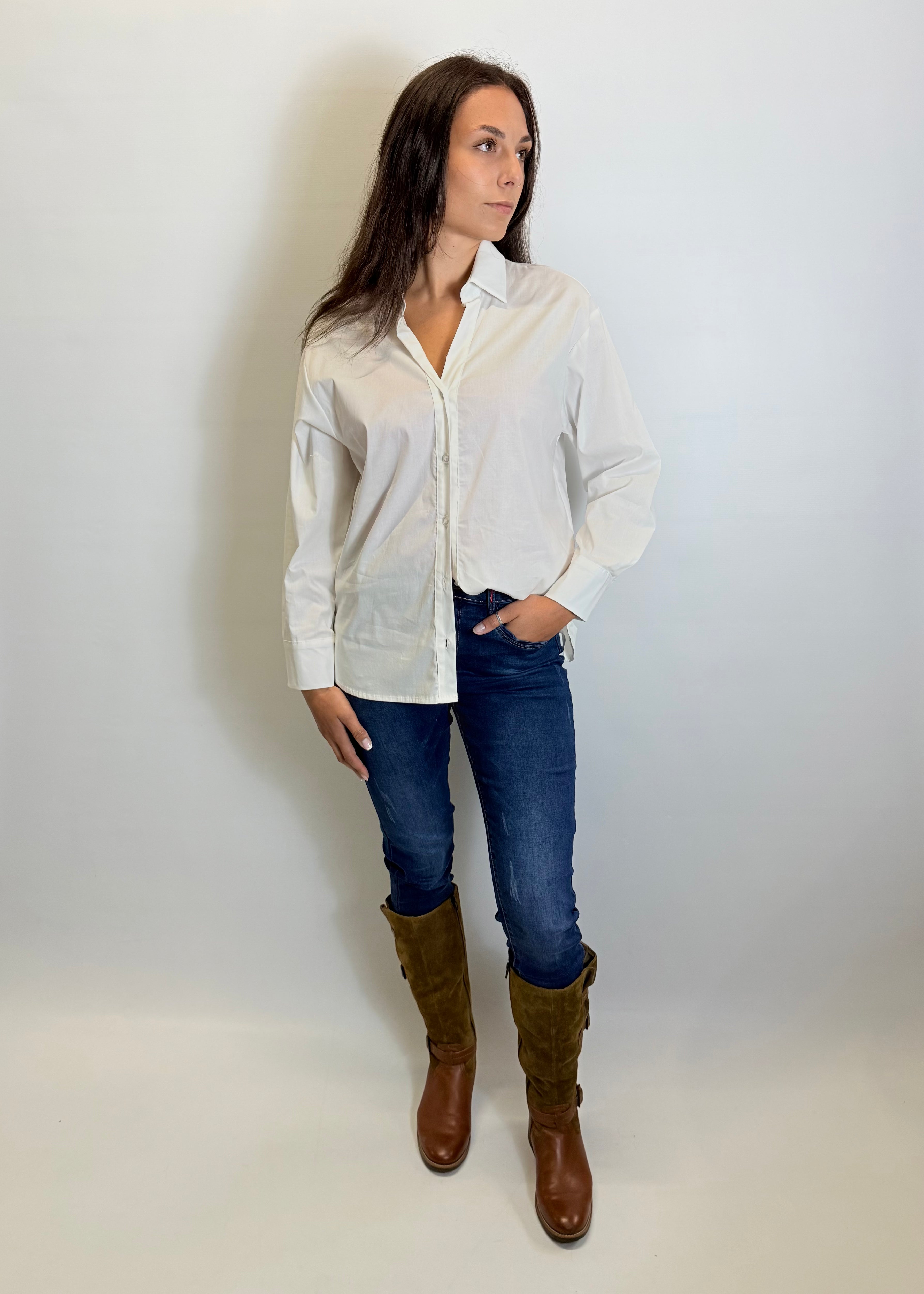 Woman wearing a white shirt, blue jeans, and brown boots against a plain background