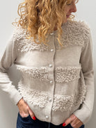 A woman with curly blonde hair wearing a beige textured knit cardigan with fuzzy trim and buttons, paired with dark blue jeans, standing against a plain light gray wall.