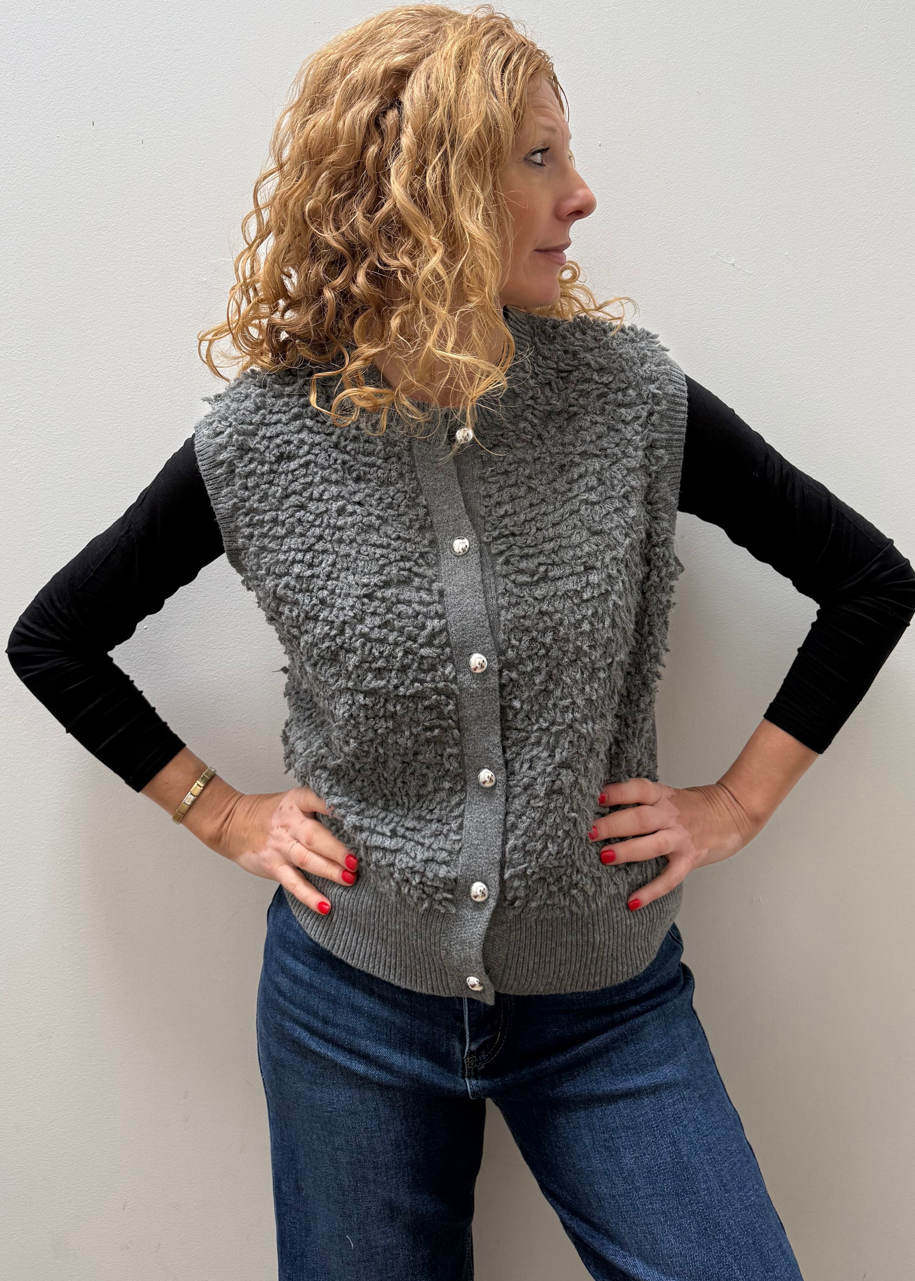 A woman with curly blonde hair models a gray textured sherpa button-up vest over a black long-sleeve shirt and blue jeans, standing against a plain light gray wall.