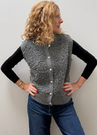 A woman with curly blonde hair models a gray textured sherpa button-up vest over a black long-sleeve shirt and blue jeans, standing against a plain light gray wall.