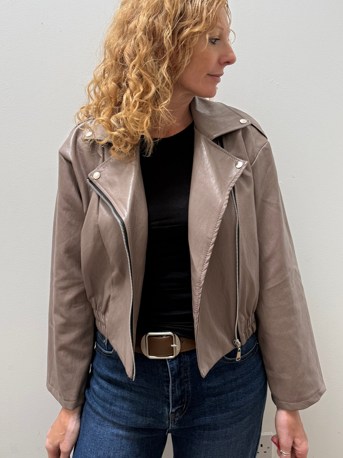 A woman with curly blonde hair wearing a taupe faux-leather moto jacket, black top, and blue jeans, standing against a plain light gray wall.