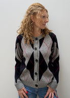 A woman with curly blonde hair wearing a gray, navy, and red argyle cardigan sweater with black buttons, paired with blue jeans, standing against a plain light gray wall.