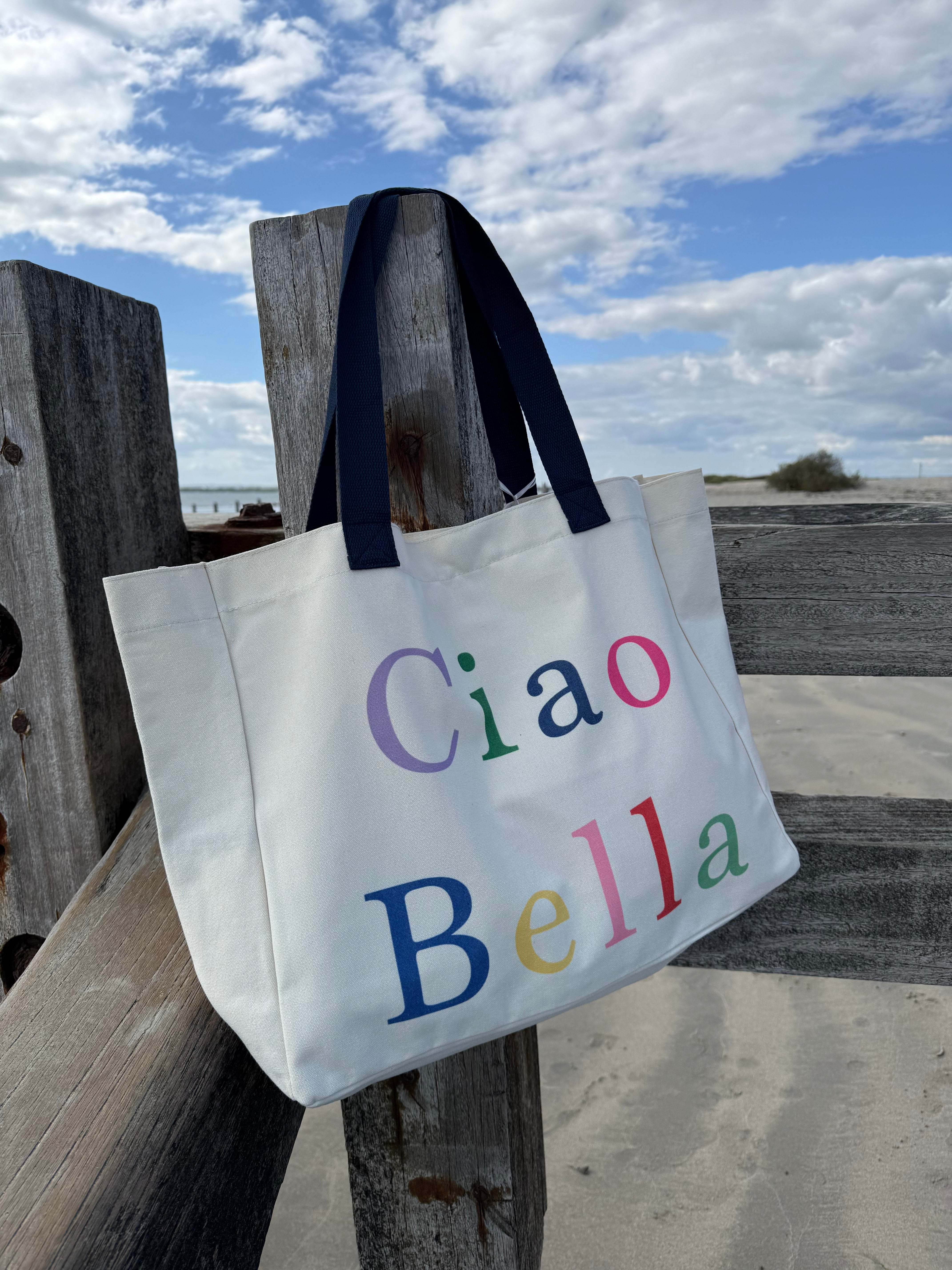 Tote bag with 'Ciao Bella' text on a wooden post against a cloudy sky.
